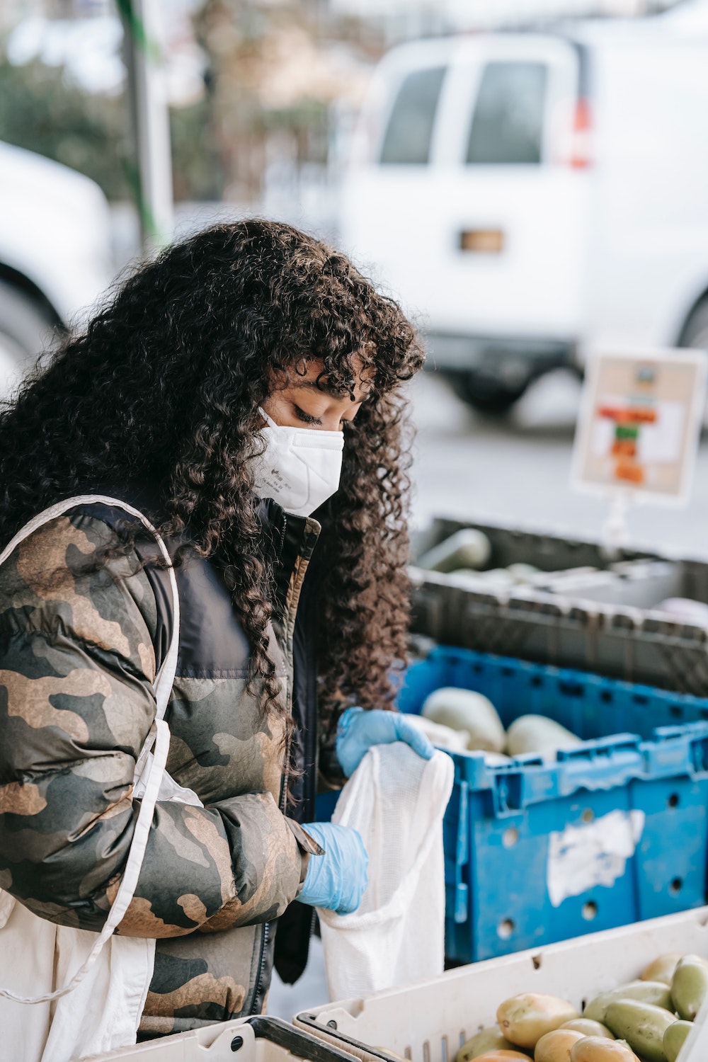 A woman is wearing a camo-patterned jacket, and a face mask. She is outdoors, buying fruits.