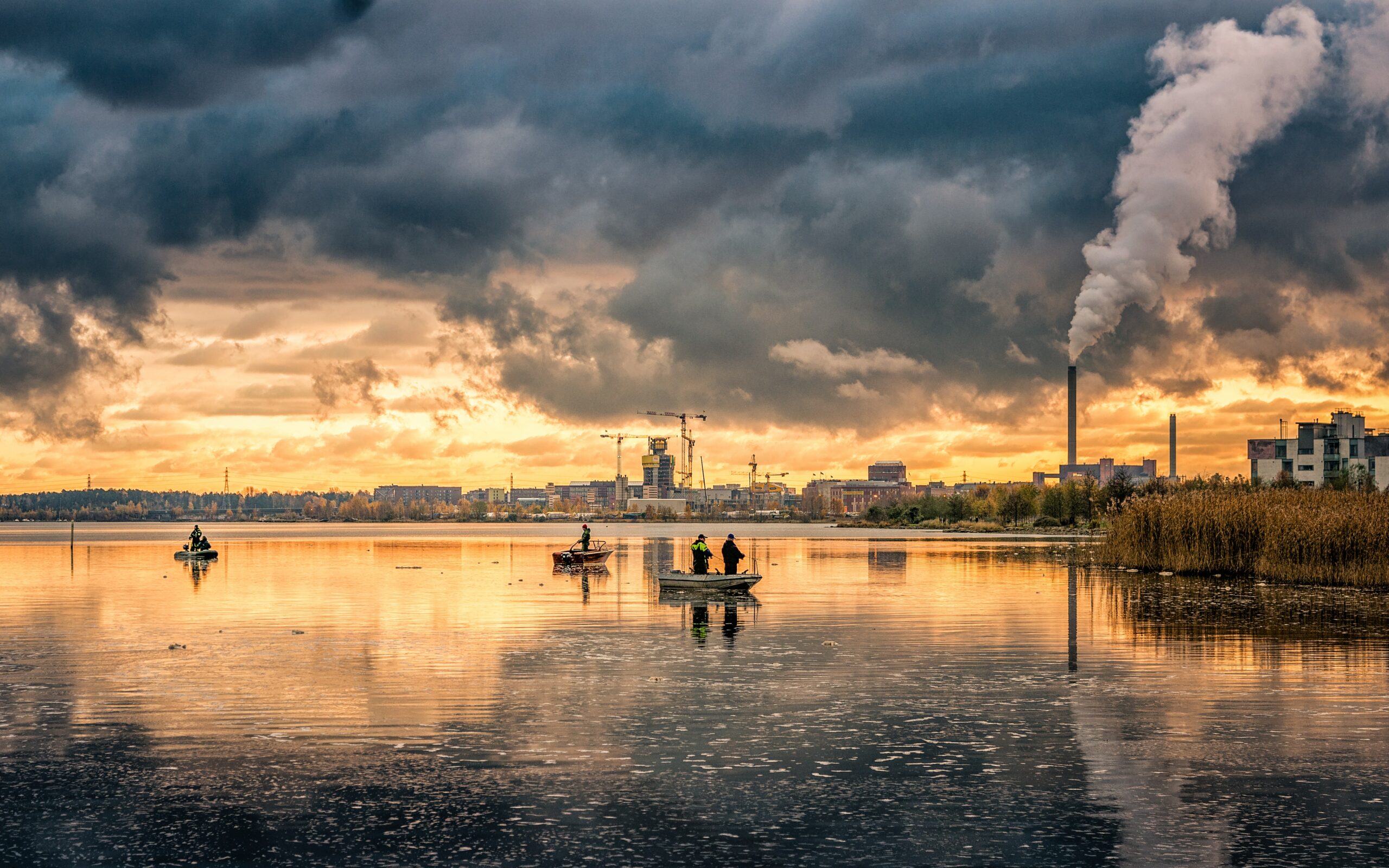 A body of water near a city. In front of an orange sky, a pillar of smoke rises from a smokestack, looking identical to the clouds overhead. People stand in small boats, fishing. The surface of the water looks speckled and dirty.