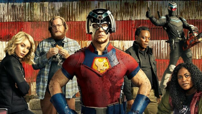 A team of DC superheroes, with John Cena as Peacemaker in the centre, posed in front of a grungey warehouse painted like an American flag