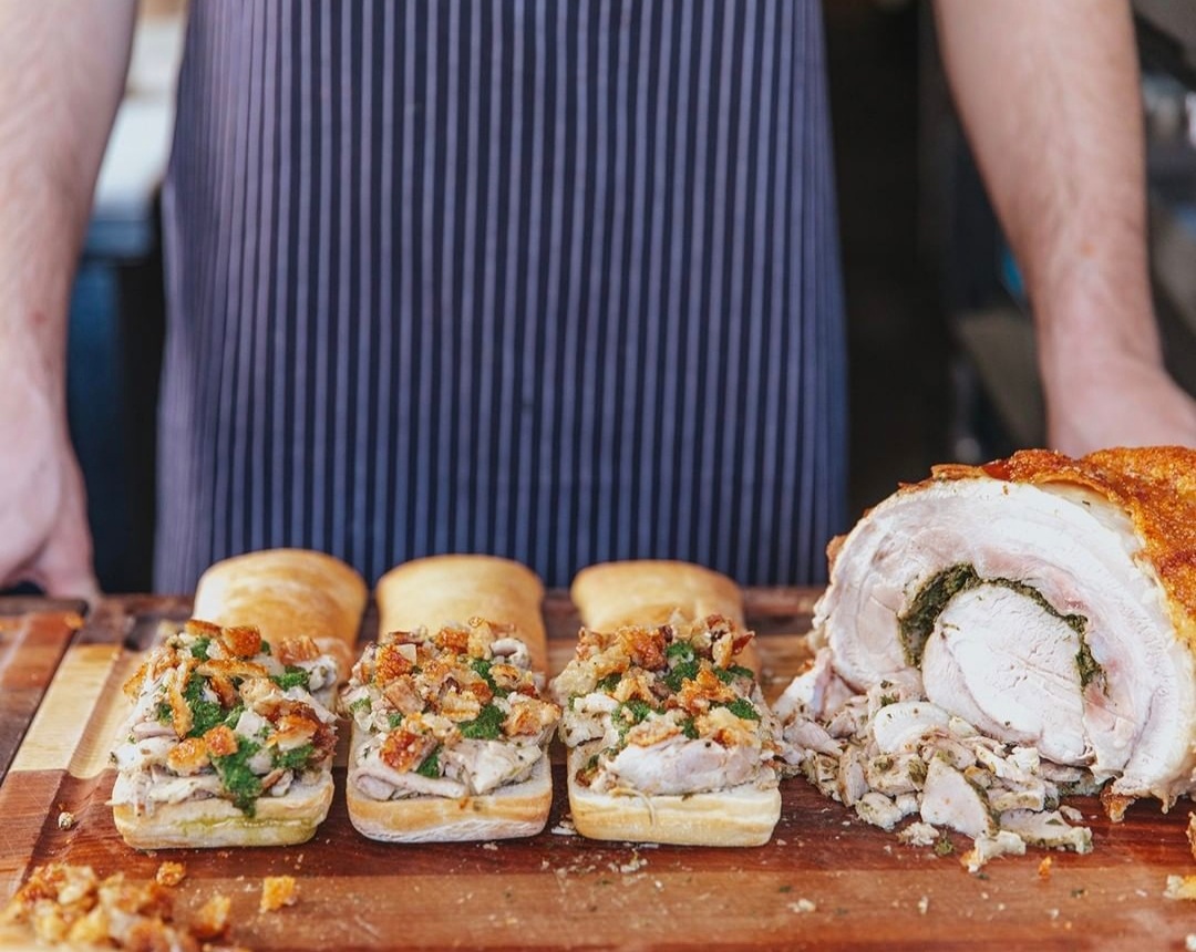 Three open face porchetta sandwiches in a row on a cutting board, with a half-carved pork roast on the right