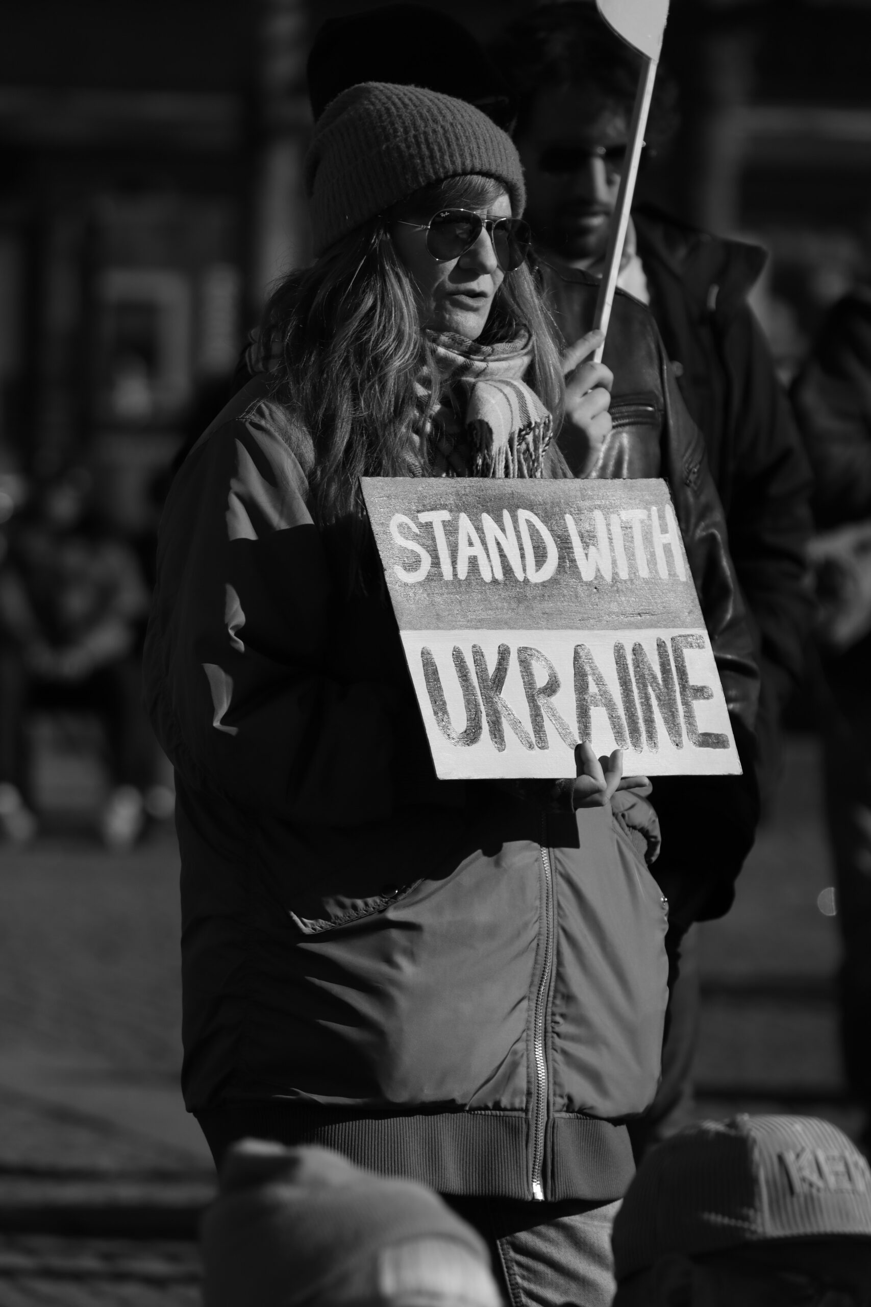 Someone is holding a sign that reads “Stand With Ukraine.” The image is in greyscale, but it looks like the sign is coloured to mirror the Ukrainian flag.