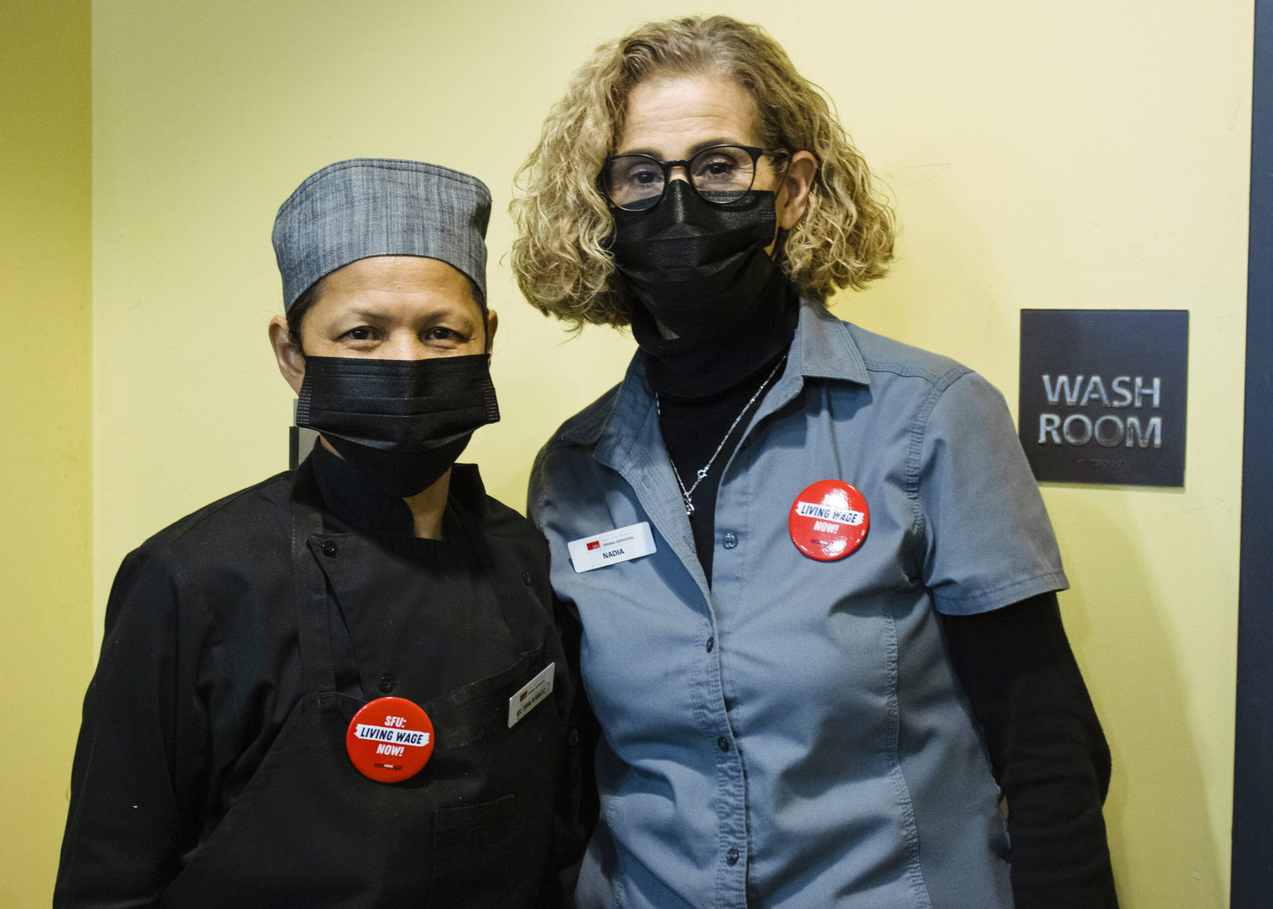 two food service workers standing next to each other, the person on the right has a red “Living Wage” pin on their uniform