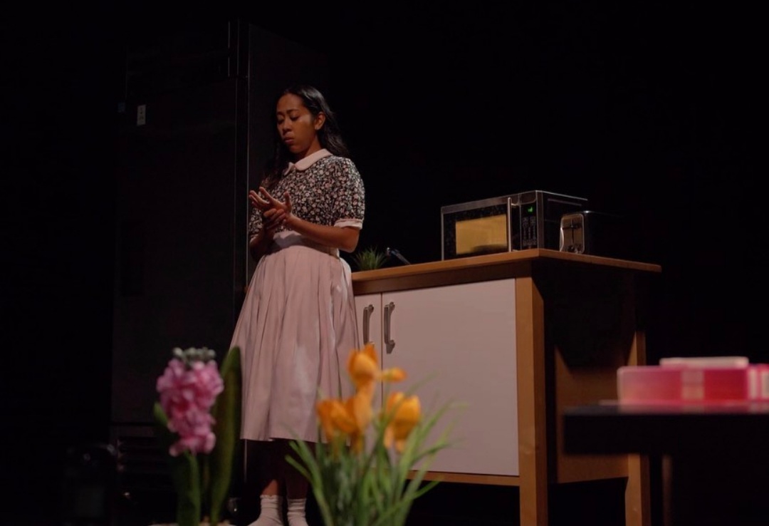 Actress Sarah Roa, dressed like a 1960s housewife, stands on stage in front of a kitchen table looking down at her hands. Pink and yellow flowers sprout up in the foreground.