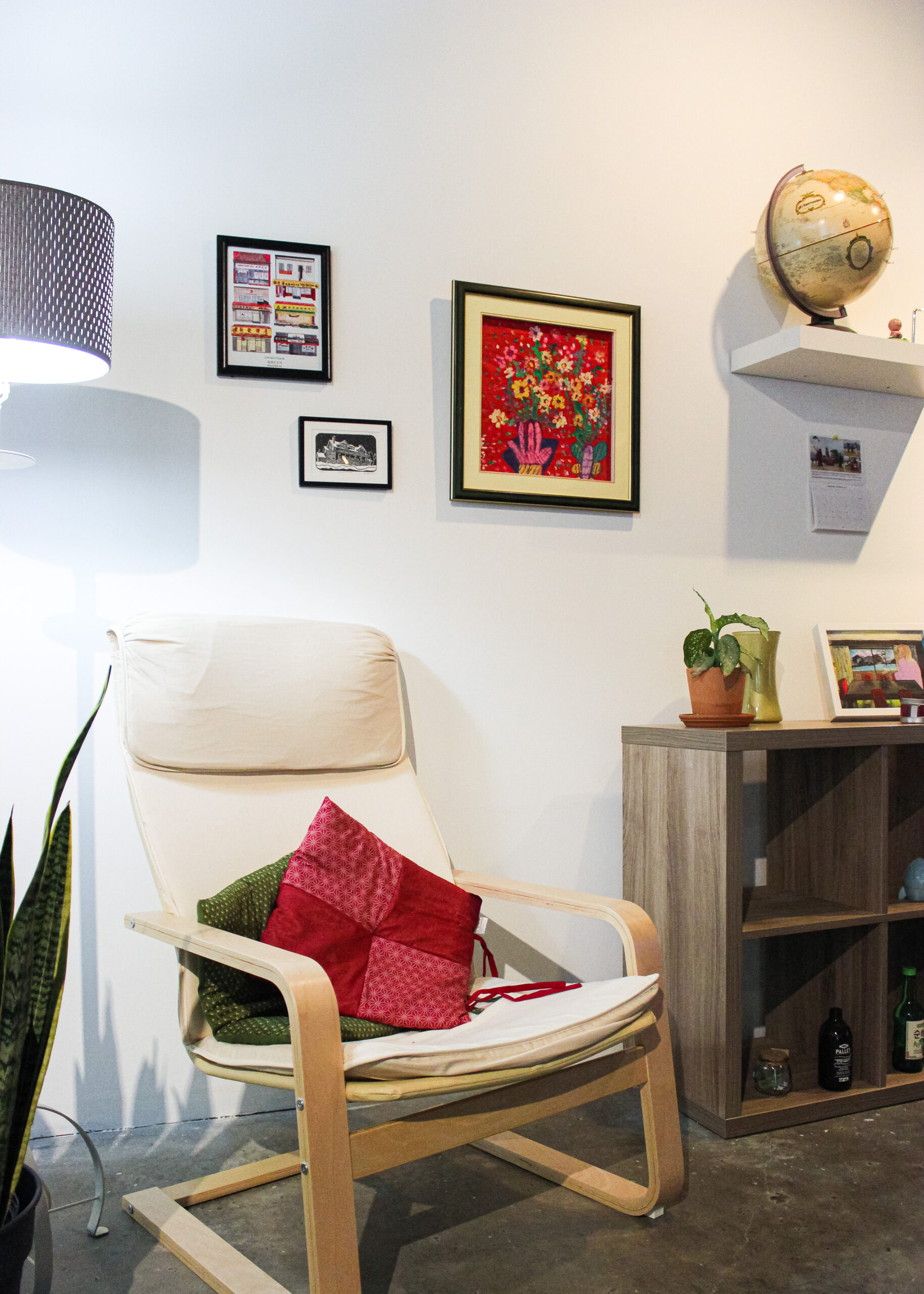 A comfy Ikea armchair against a white wall with framed art prints. To the right side, a storage unit with plants and bottles of sake. To the left, a floor lamp turned on.