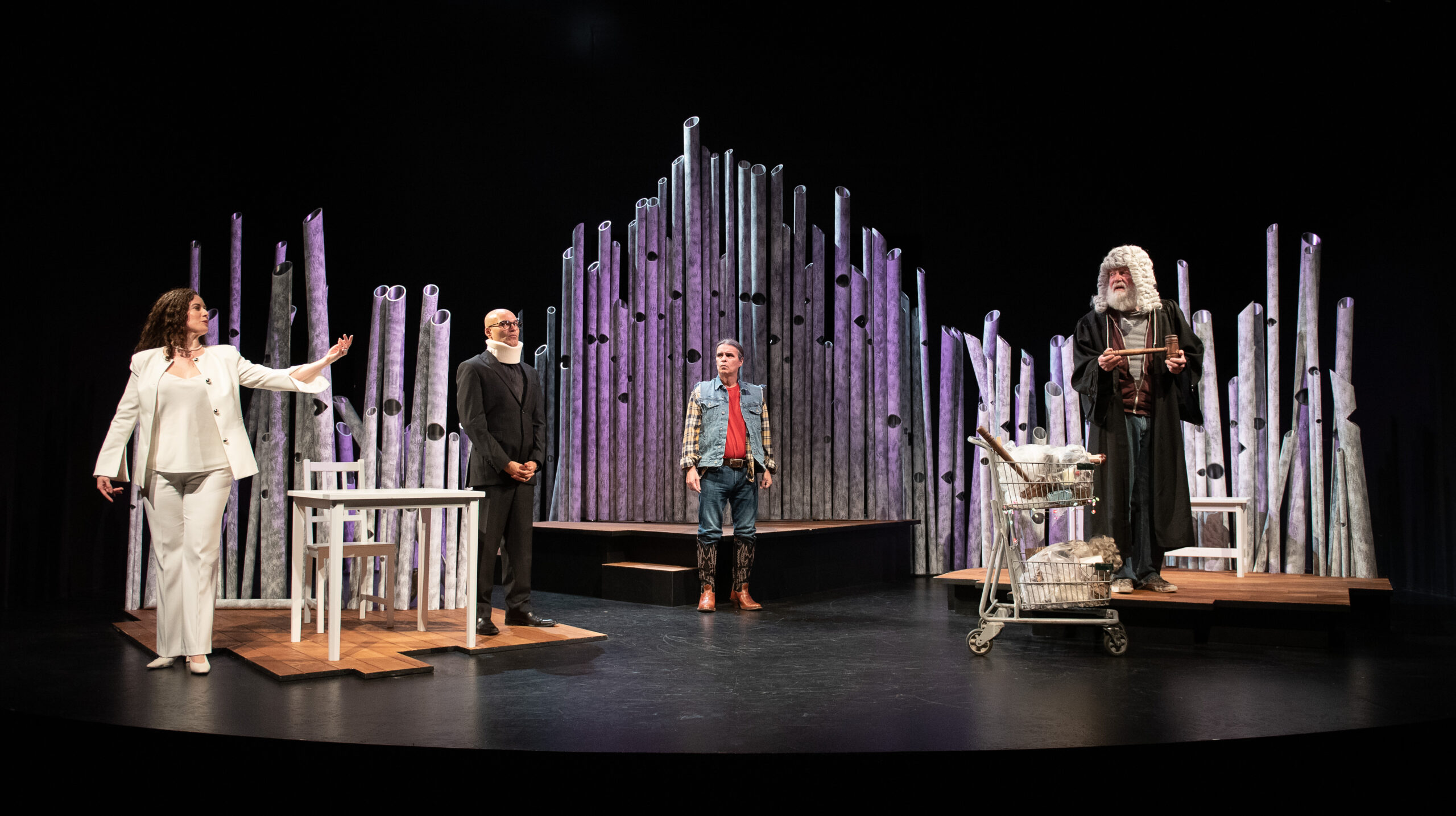 A curved stage featuring a dead forest set design in the background. In the spotlight, from stage right: a woman in a white power suit, a bald man with a neck brace, the Little Red Warrior dressed like a lumberjack, and a judge