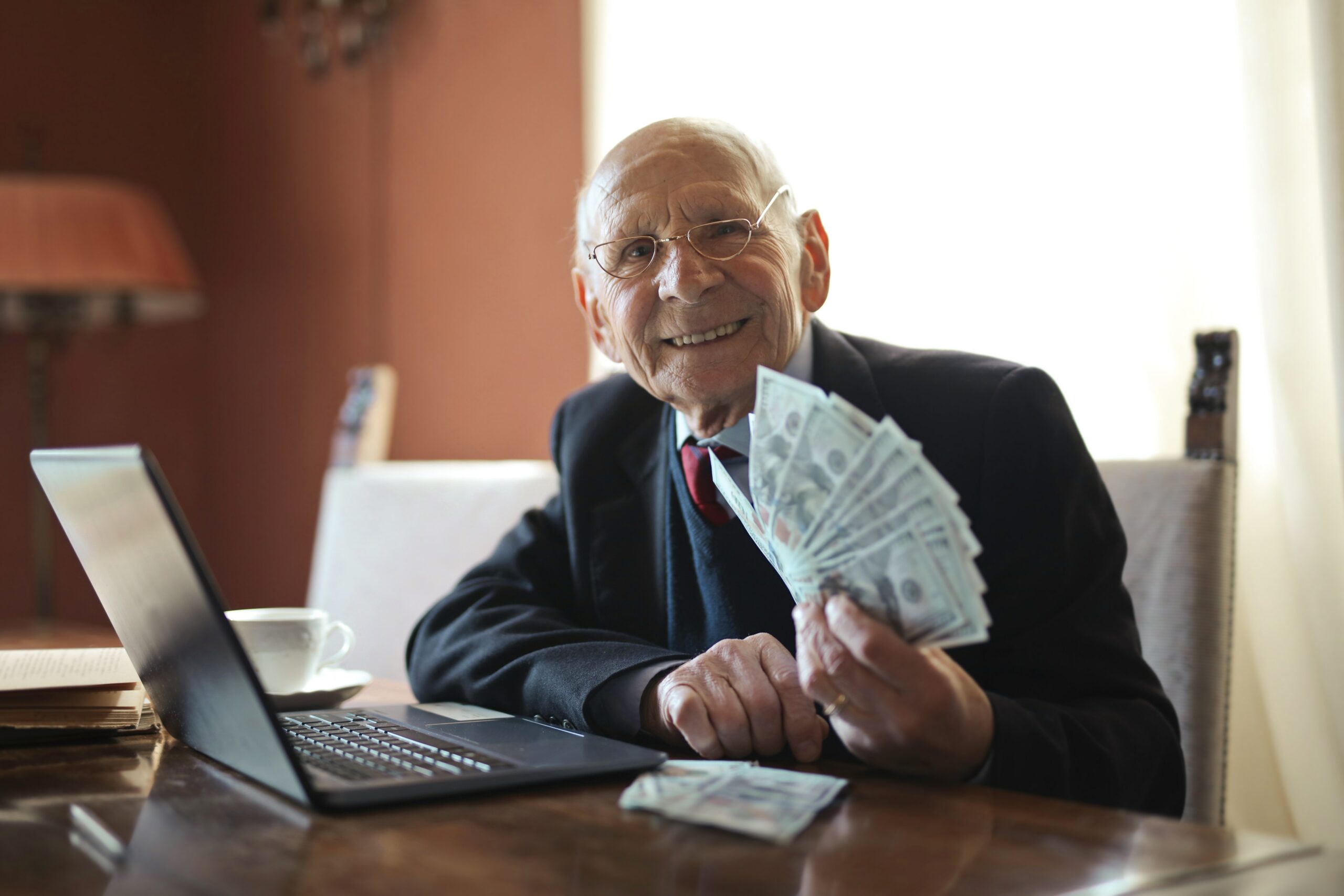 An aged white man in a suit sits in front of a laptop. He is staring at the camera, and is fanning out a large number of bills in his hand.