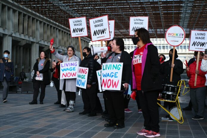 SFU workers are seen at SFU holding signs that read “Living Wage Now!” There are 11 people in the photo, most holding signs. One holds a megaphone and appears to be speaking.