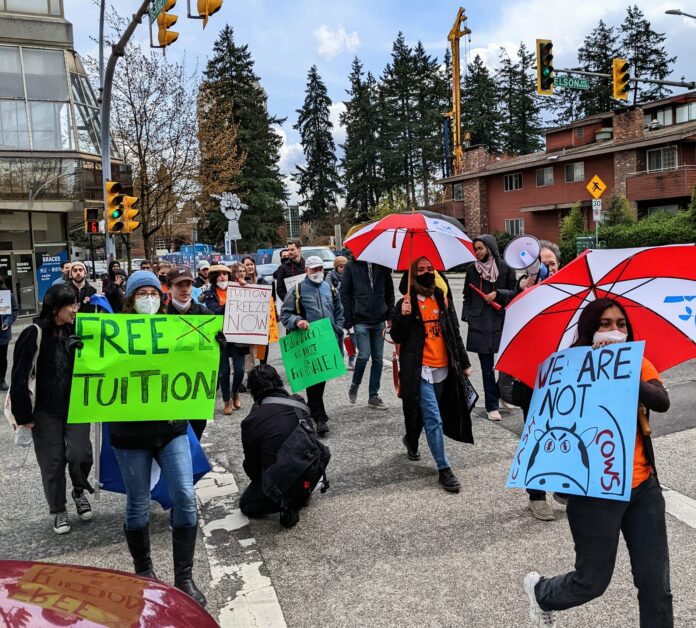 Sude Guvendik discusses the toll tuition increases have on international students. Image courtesy of Sima Jamali. Numerous protestors are seen crossing the street. Most are holding signs that read “Free Tuition” and “We Are Not Cash Cows.” Others hold umbrellas while one individual is holding a megaphone.