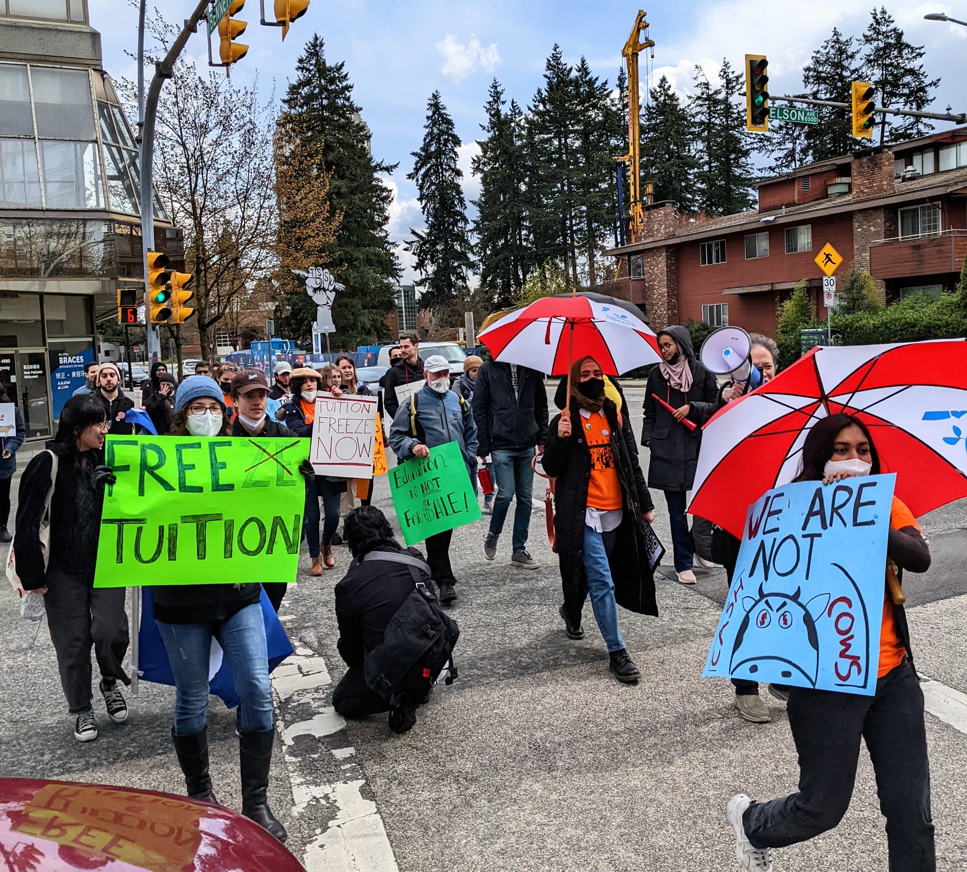 Numerous protestors are seen crossing the street. Most are holding signs that read “Free Tuition” and “We Are Not Cash Cows.” Others hold umbrellas while one individual is holding a megaphone.
