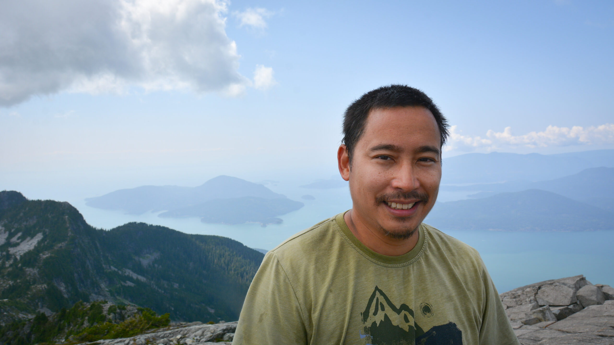 Stephen Hui is looking into the camera smiling. He is standing atop Twin Sisters. Mountains, water, and a blue sky is behind him.