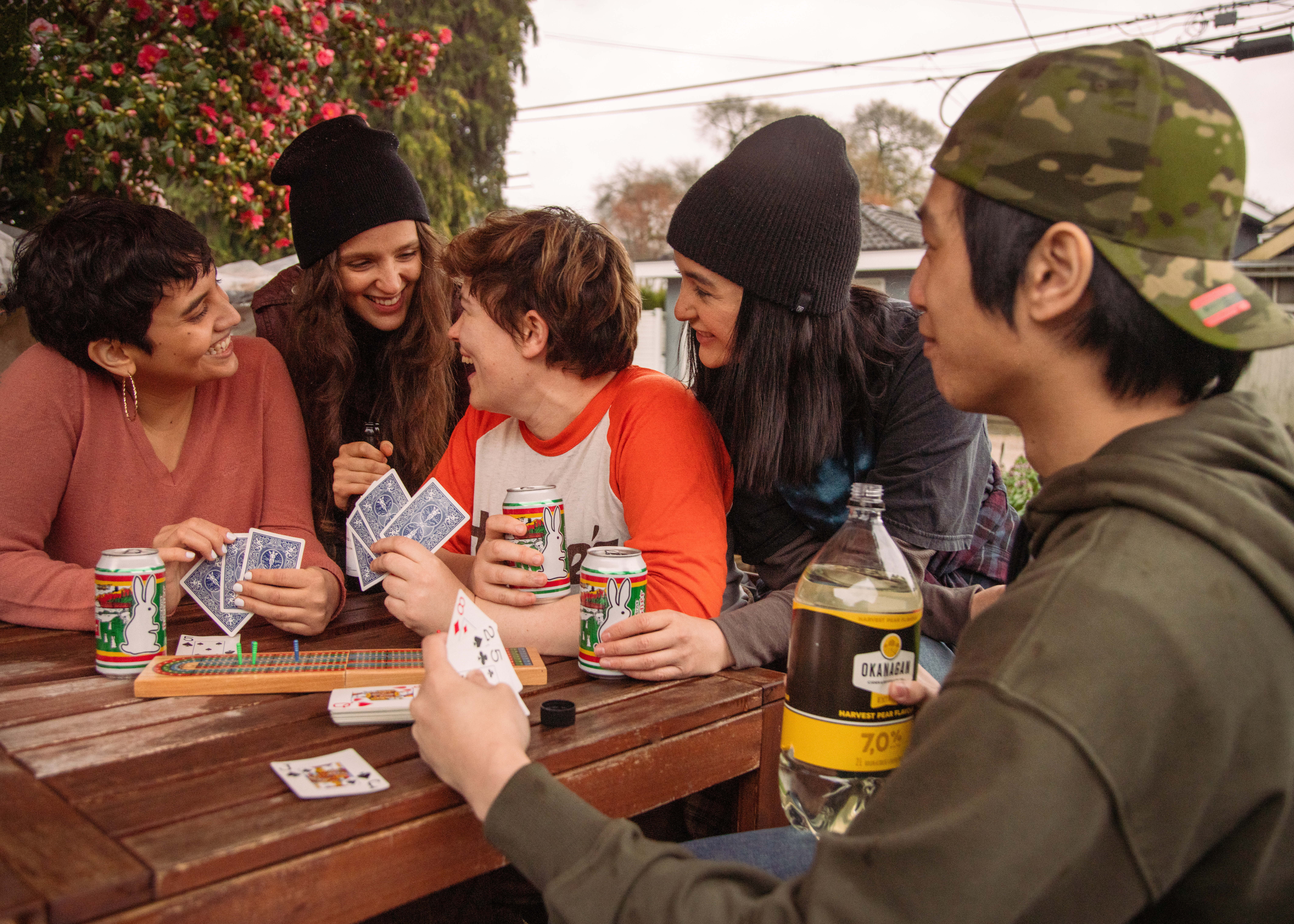 Five young-adult friends sit around a small outdoor table with beers and playing cards in hand