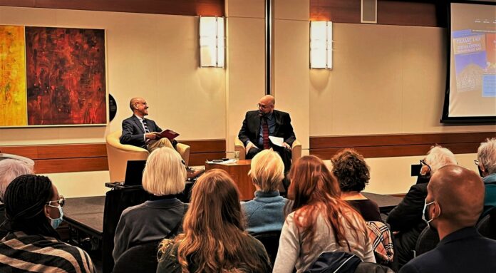 Dr. Emon is a professor of law and history at University of Toronto. Image courtesy of Nabila Hussein Dr. Anver Emon is seen sitting on stage with Dr. Amyn Sajoo. The two are engaged in conversation as people sit in the audience listening. Behind the stage are paintings and lights.