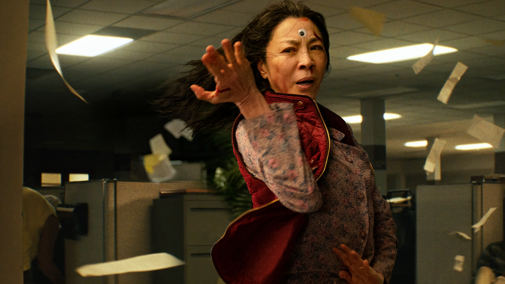 Action shot of middle-aged Chinese woman in a floral shirt and red vest, twisting her arm as though in a fighting position. Background is a darkened office with papers flying around her.