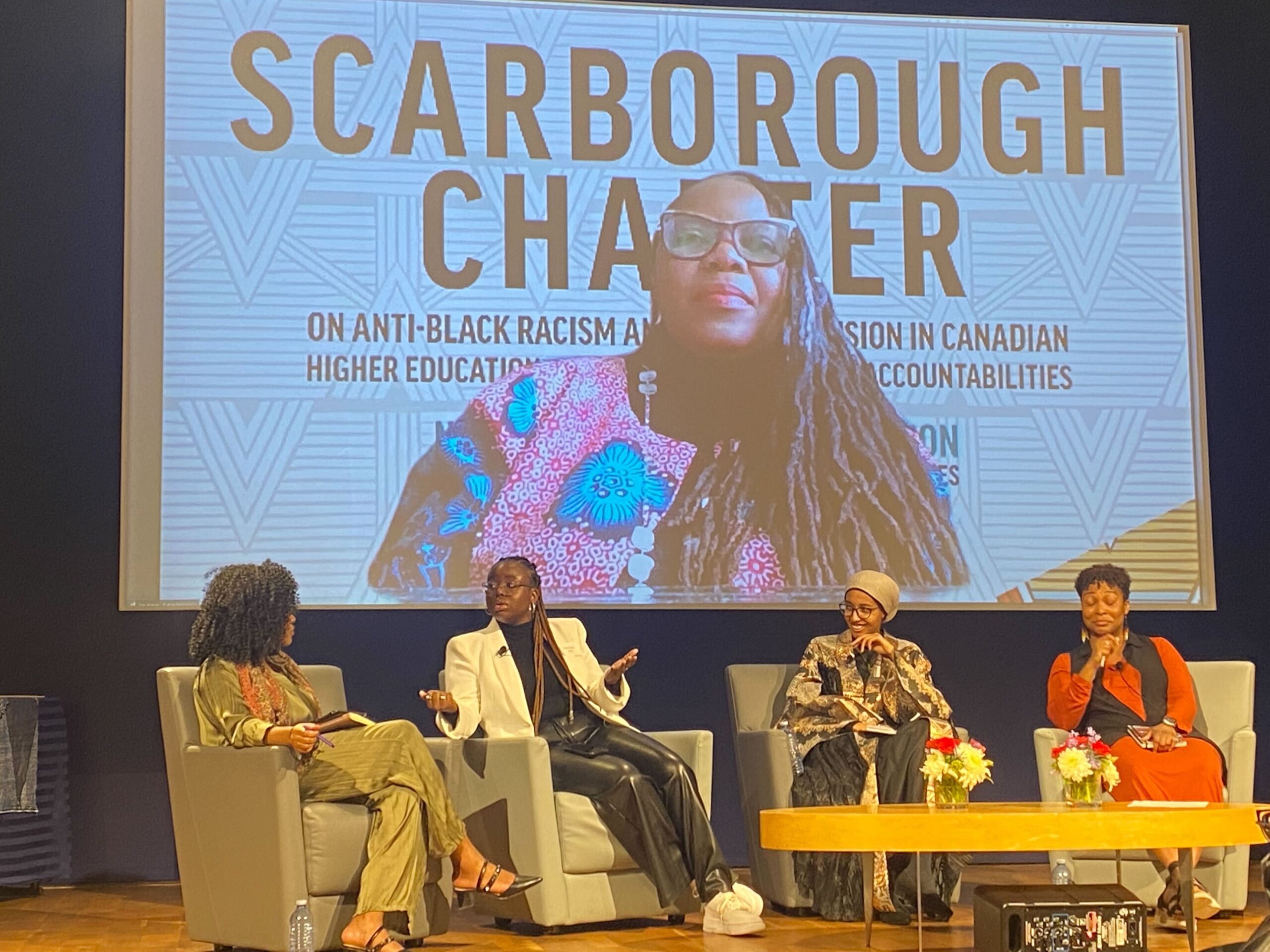 The photo is of four Black panellists engaging in conversation onstage. Behind them is a screen that reads Scarborough charter. The screen also has another panelist joining in virtually.