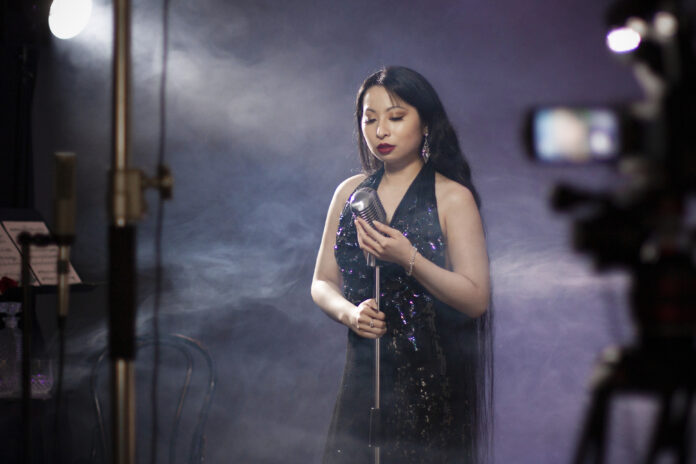 JacquelineKo_BehindTheScenes_PhotoByStephanieKo_PRINT Singer Jacqueline Ko is standing in a sequined dress in front of a vintage microphone. She is spotlighted by a beam of light, against a dark backdrop of purple mist.