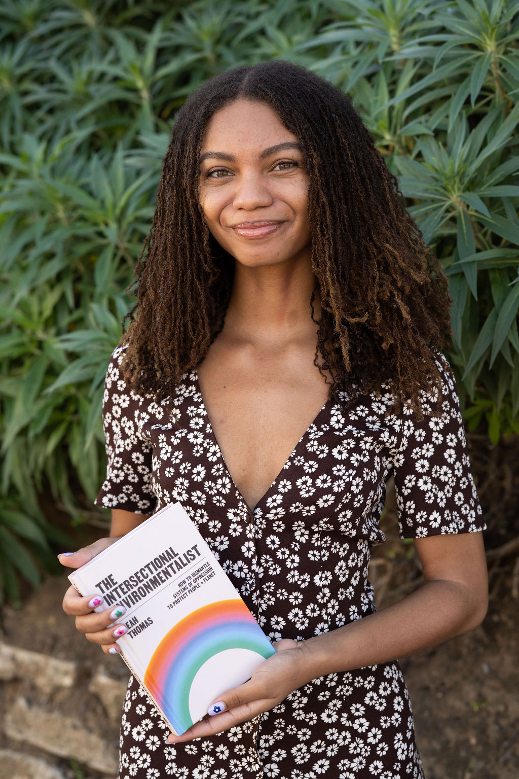 Leah Thomas is standing, looking into the camera, while holding her book, the intersectional environmentalist. She is standing in front of a wall of large green plants.
