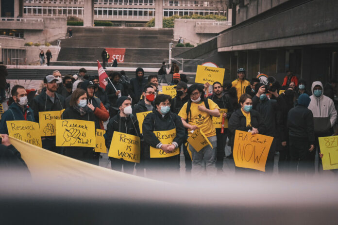 Day 873 of SFU’s delay in bargaining. Image courtesy of Sherry Young. A crowd of people can be seen at SFU Convocation Mall. They’re holding yellow signs that read “Research is Work” and “RA Contract Now”.