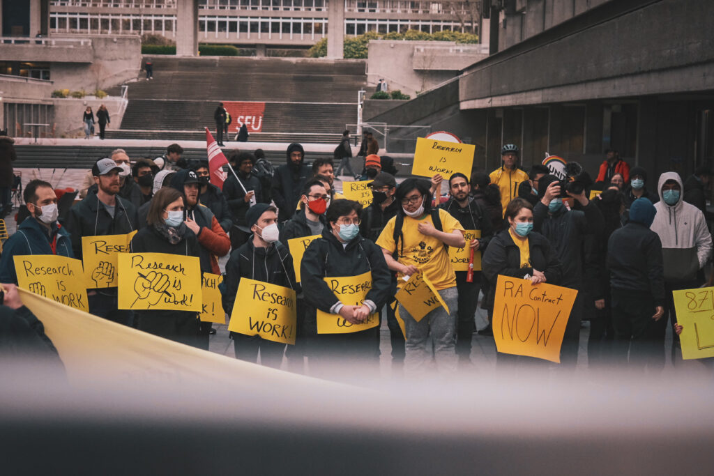 A crowd of people can be seen at SFU Convocation Mall. They’re holding yellow signs that read “Research is Work” and “RA Contract Now”.