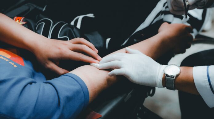 blood an individual in a blue shirt and black pants is sitting down with his arm outstretched. A doctor wearing white gloves is applying pressure to his arm with gauze. The individual is donating blood.