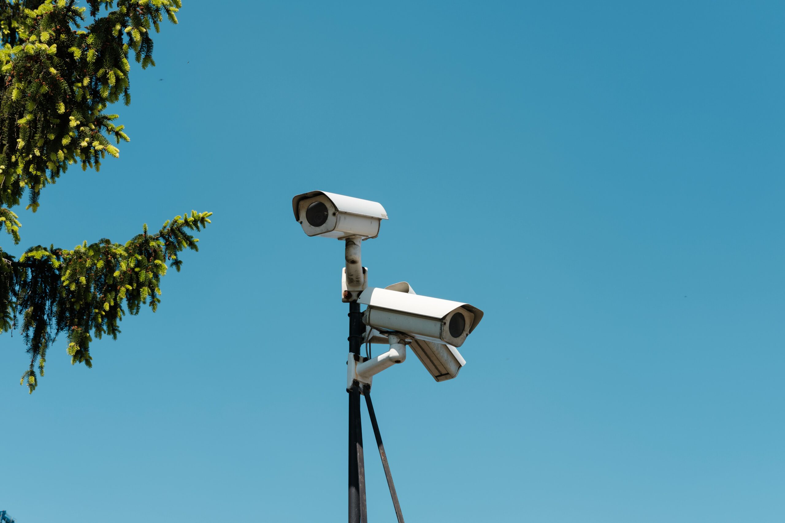 Two CCTV cameras are seen on a pole. Their background is a bright blue sky.