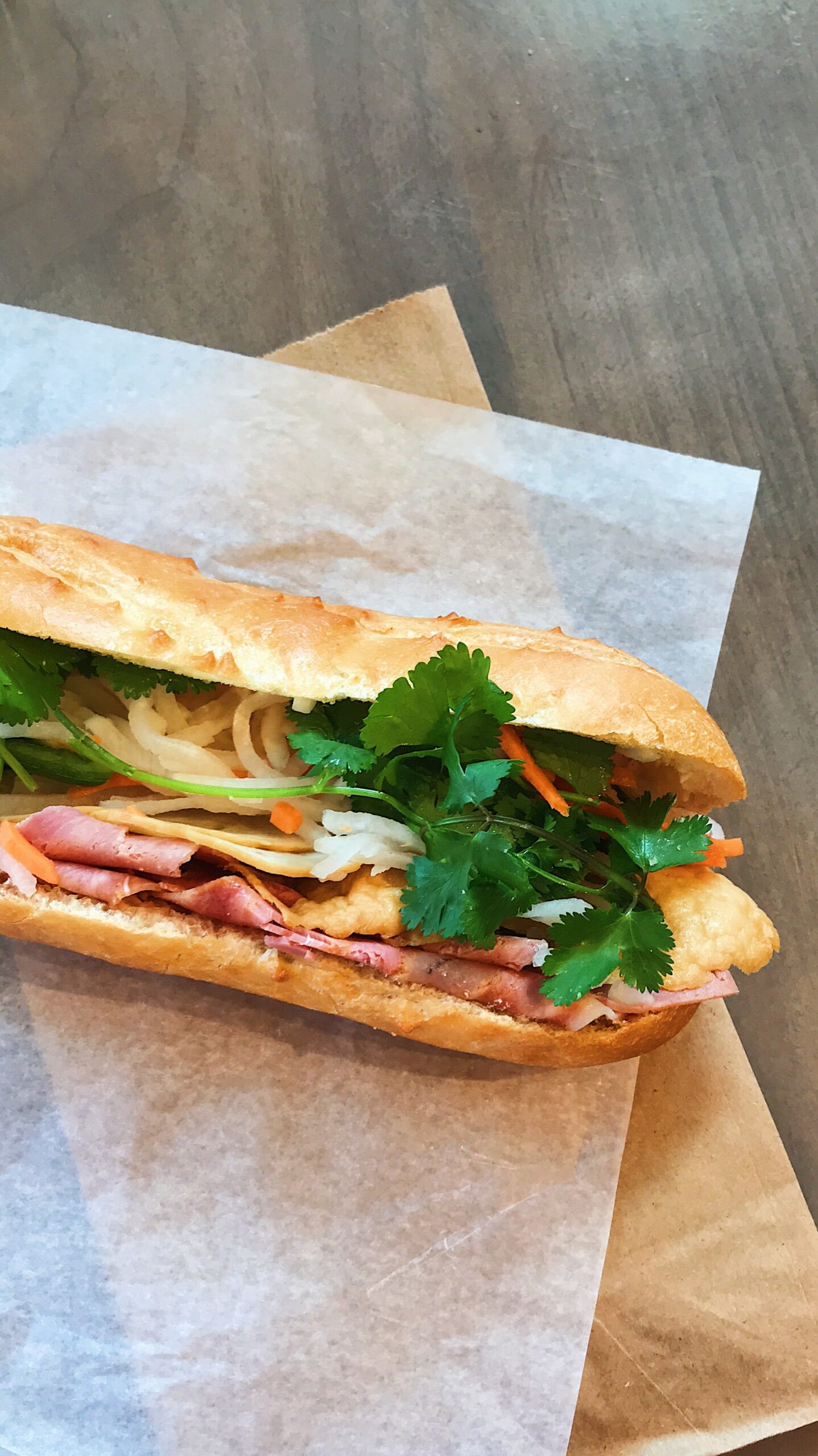 Photo of a bánh mì sandwich on a sheet of parchment paper and a paper bag beneath it.