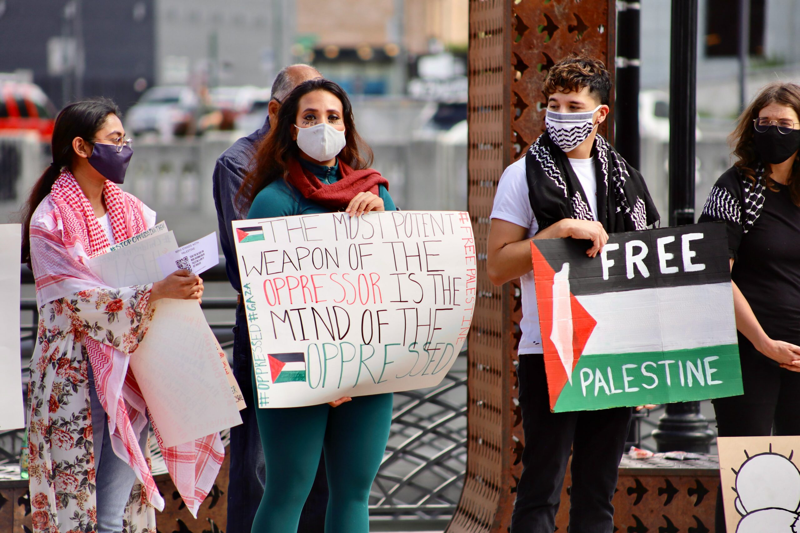 Protestors are standing holding signs that read “Free Palestine” and “The most potent weapon of the oppressor is the mind of the oppressed.” An individual in all blue is staring at the camera, while those around them are looking in their direction.