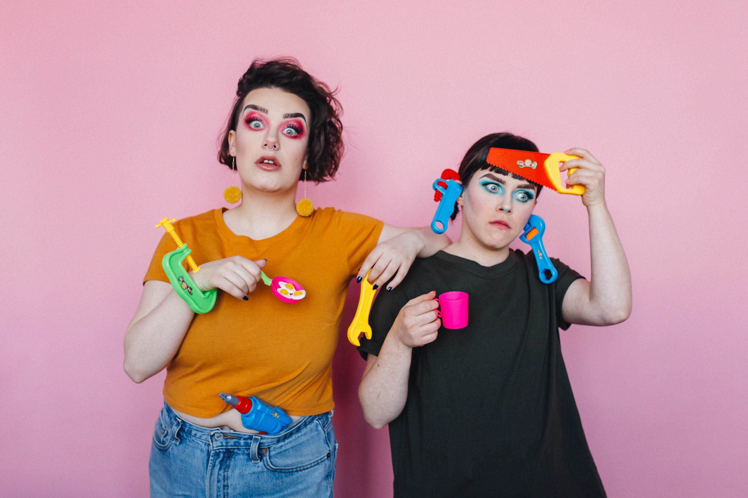 Two individuals standing against a pink background armed with various children’s toy construction tools. They wear shocked and confused facial expressions and blue and pink eyeshadow.