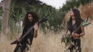 Two women dressed in all-black play electric guitars in the midst of tall grass