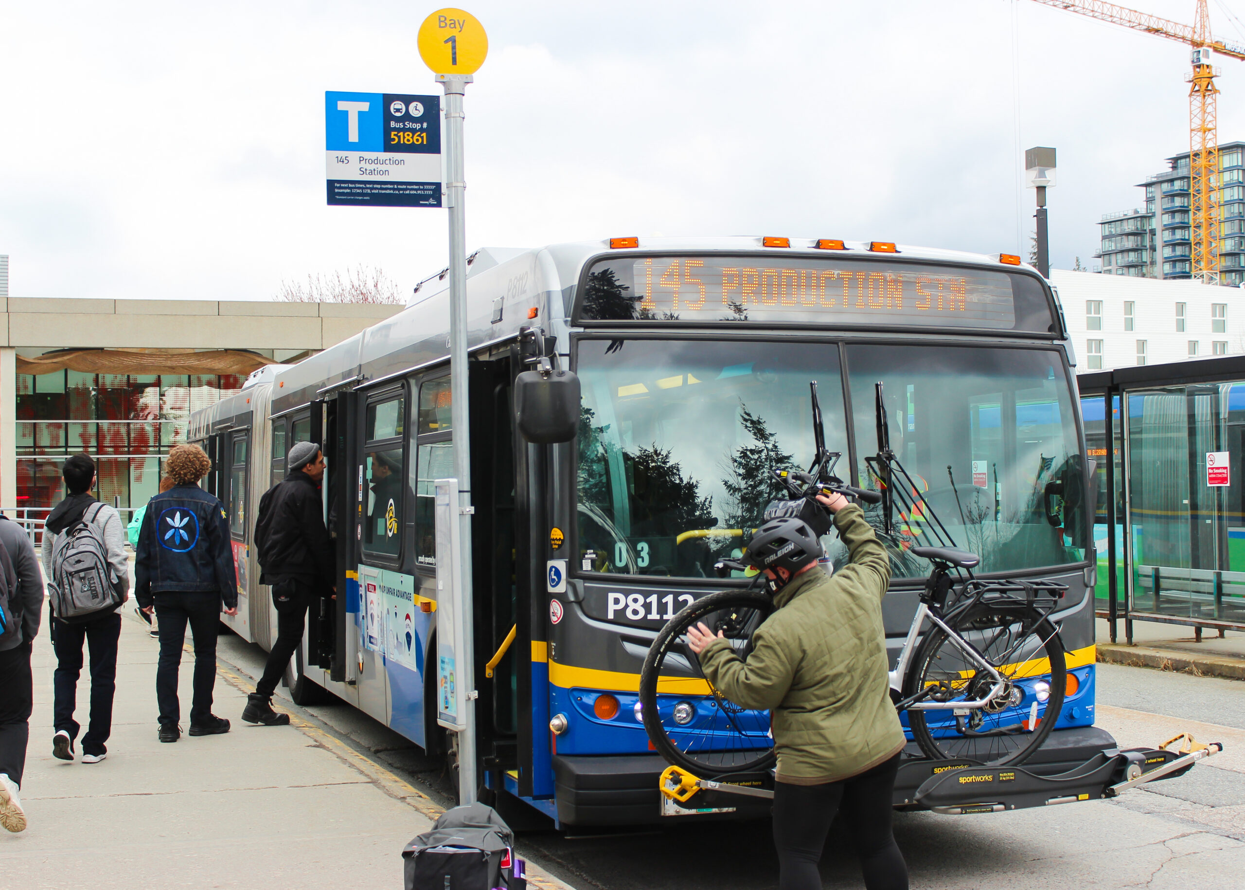 The photo shows the front of a BC TransLink bus. An individual is loading their bike to the front of the bus, as other passengers are boarding.
