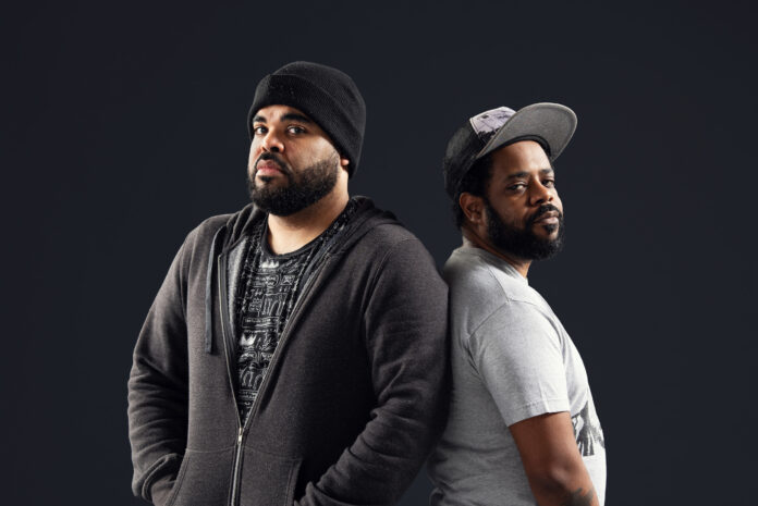 Two young Black men dressed in casual street clothes pose standing with the backs of one shoulder connected, looking at the camera solemnly