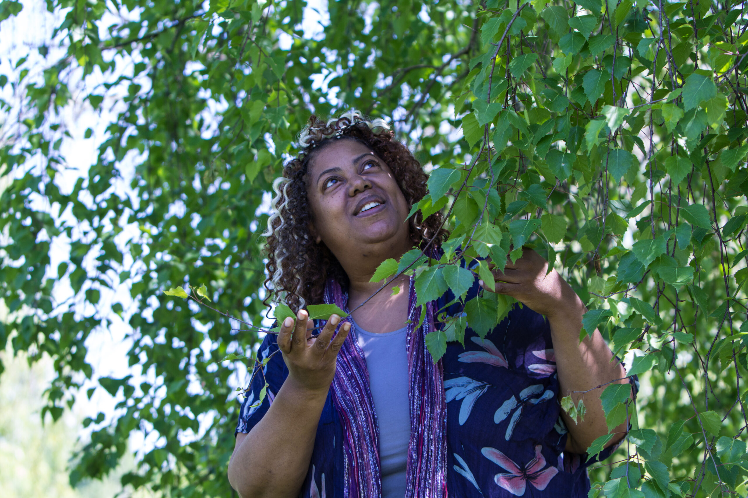 Siobhan Barker wearing a blue outfit, stands under a tree holding one of its branches full of green leaves gently, smiling upwards.