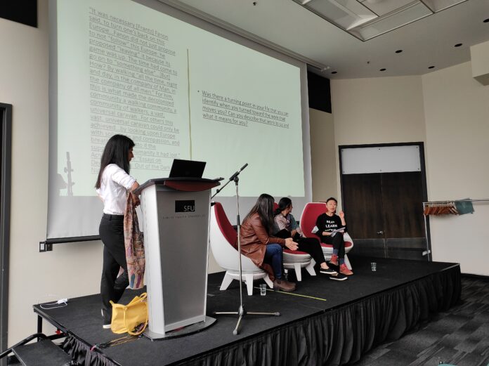 anti-asian racism event The photo is of three speakers sitting on stage, while the moderator stands next to them by the podium. Behind them is a screen with a slideshow playing.