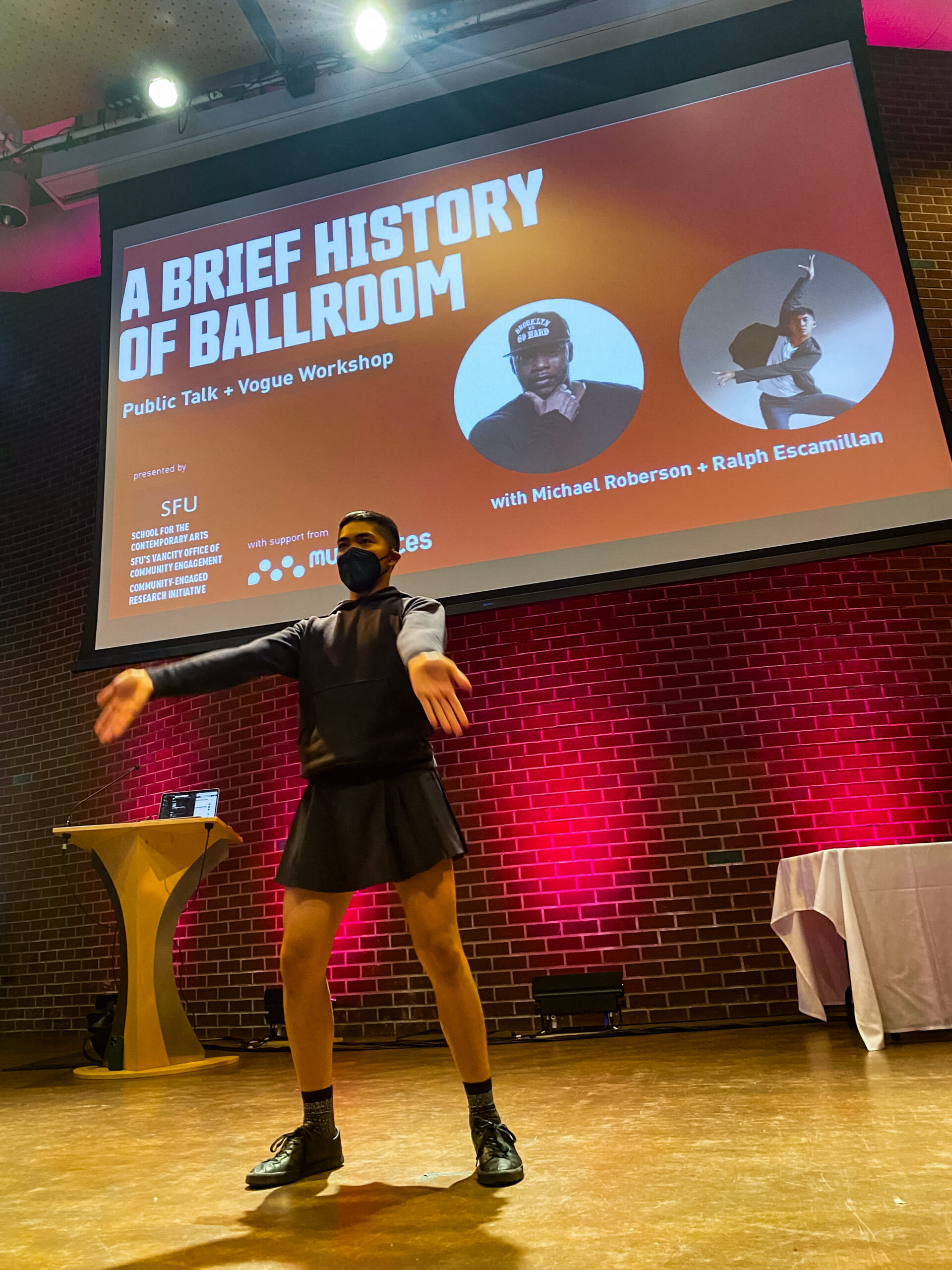 The person extends their arms out toward the audience in a vogue pose with a PowerPoint slide of the event banner projected behind him