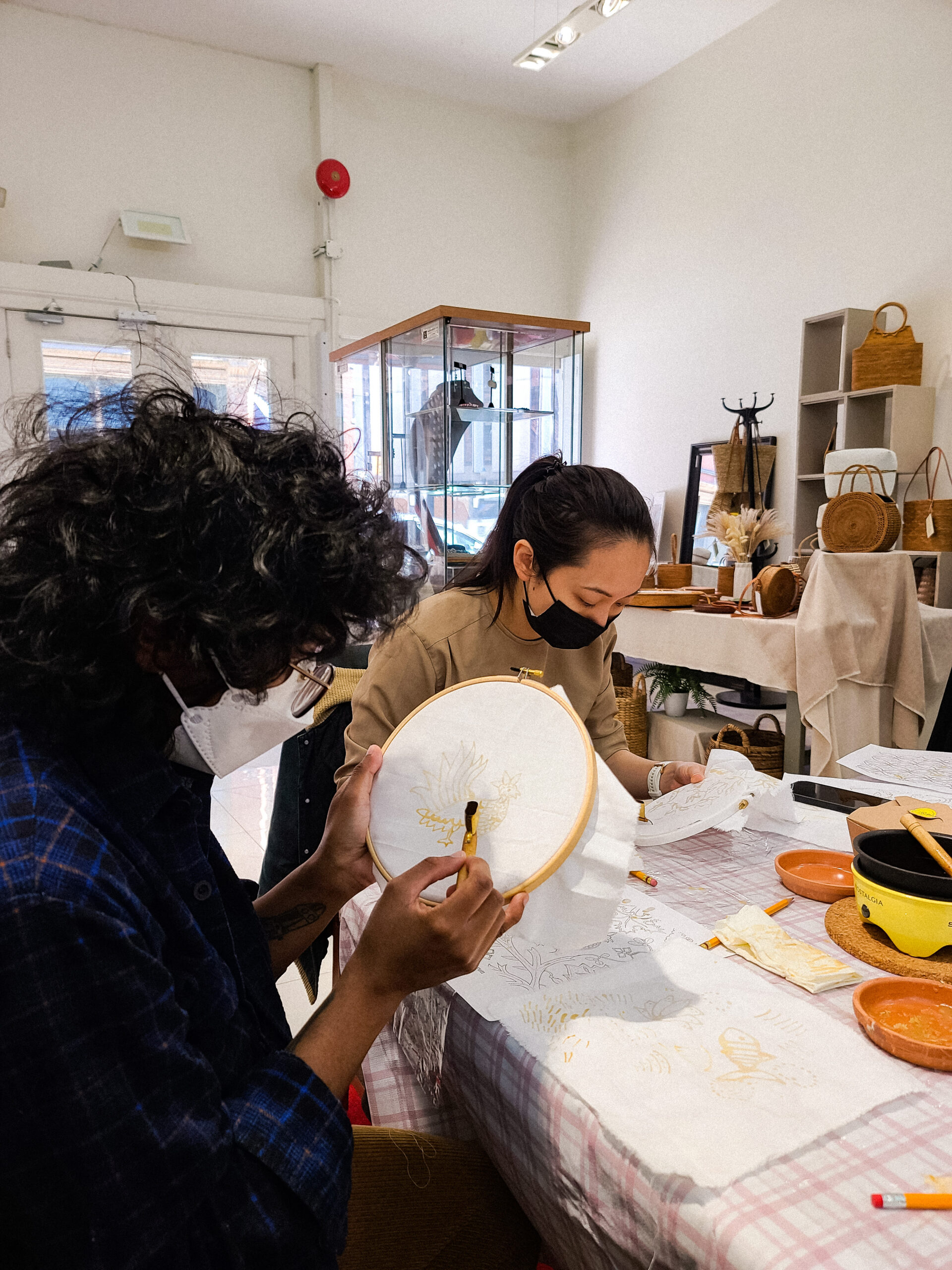 (1) Image of a person neck down wearing a moss green crewneck, holding up an embroidery hoop and using a Batik tool to draw on the fabric with wax. (2) first-person point of view, two pieces of cloth flatlaying on a table with designs drawn on them with wax. One is a symmetrical pattern that evolves into two small faces as it goes down, and one drawing is a larger side profile of a face. The person taking the photo has one hand in the photo holding up a Batik tool over the fabrics. (3) Person with dark curly shoulder-length hair, glasses, and a blue plaid shirt on has shoulders angled towards an embroidery hoop they are holding up and drawing on using a Batik tool. In the background, another person with a ponytail and moss green crewneck is seen at the table also hunched over their own Batik piece.