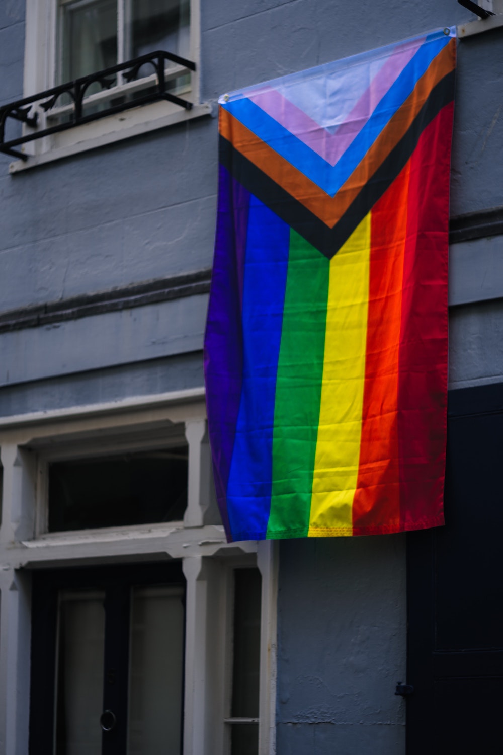 A pride flag hangs in front of a grey building. The flag includes the rainbow colours, as well as the pink, blue, brown, and black stripes.