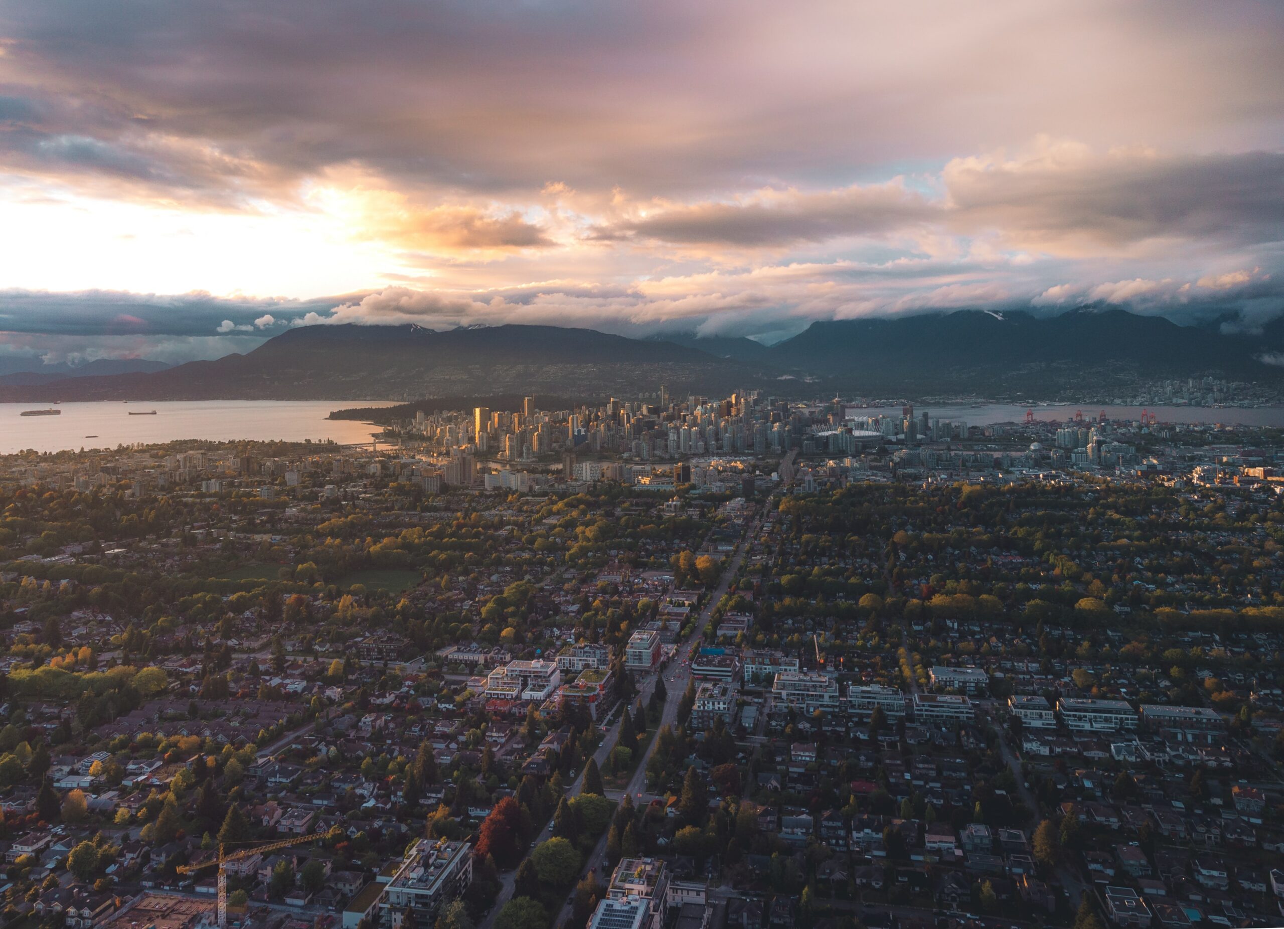 The photo shows the city of Vancouver from an aerial view. The city is in front of mountains and surrounded by water. The suburbs of Vancouver stretch towards the camera.