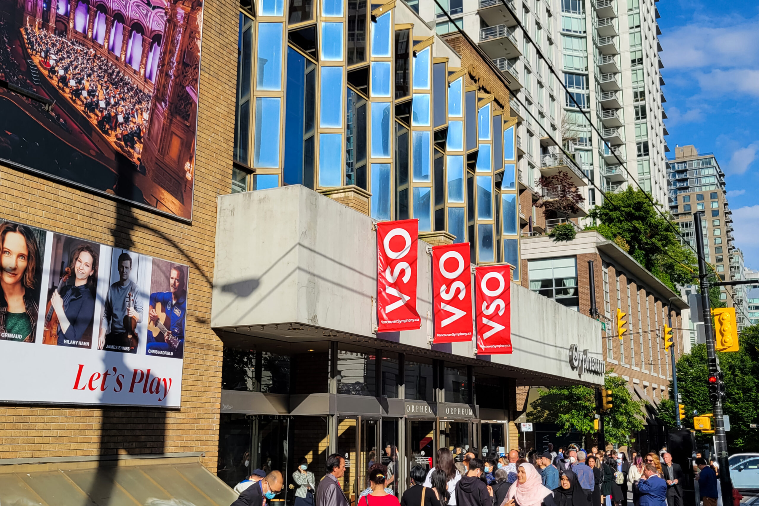 Entrance of the Orpheum with red “VSO” banners and orchestra posters hanging on the wall of the building, a fair amount of people are shown crowded at the entrance.