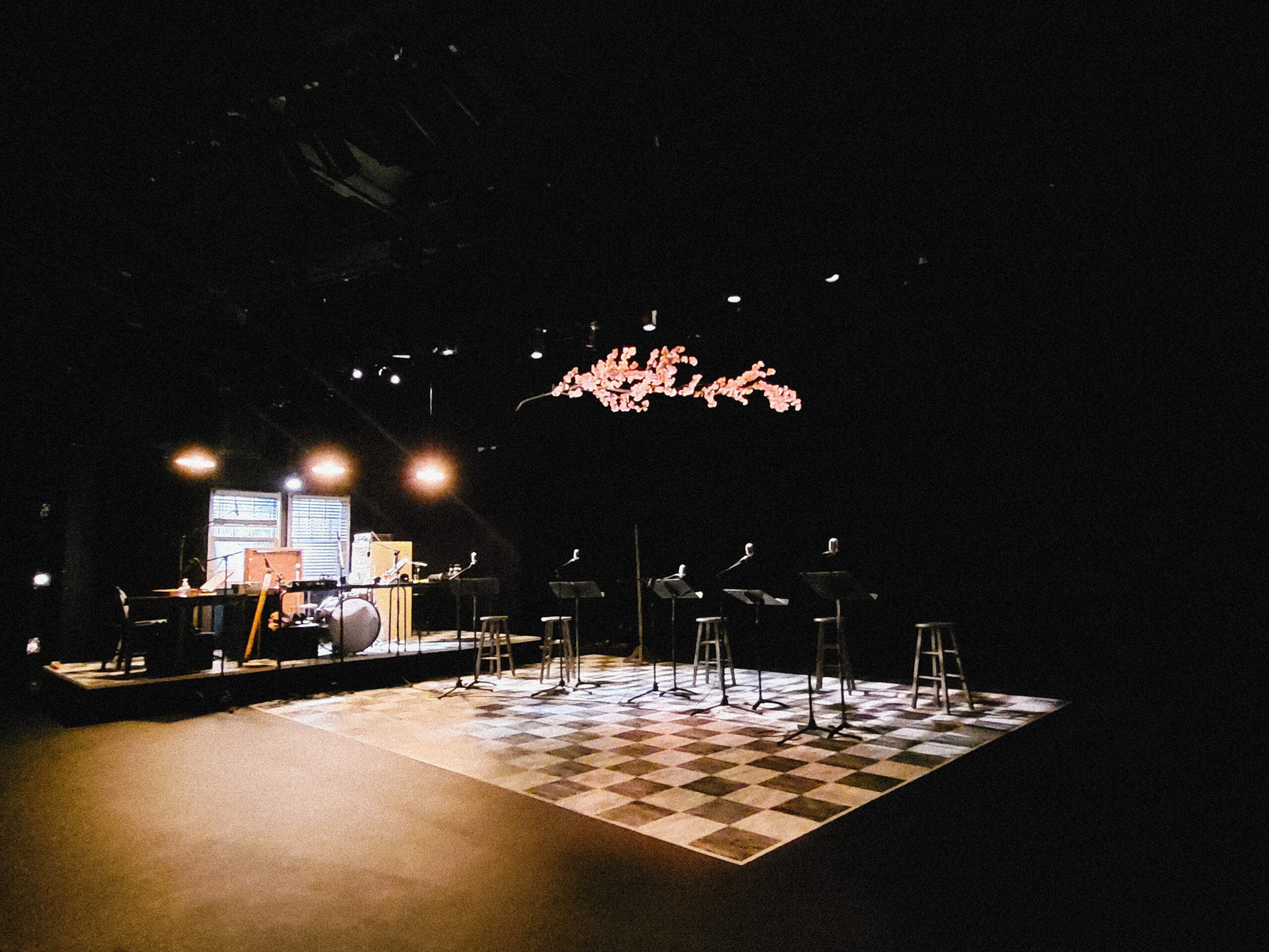 Darkened stage set with warm spotlights cast on a checkered floor decal with 5 stools set up for a Q&A. Behind them is a glowing projection of a pink cherry blossom branch extending out horizontally.