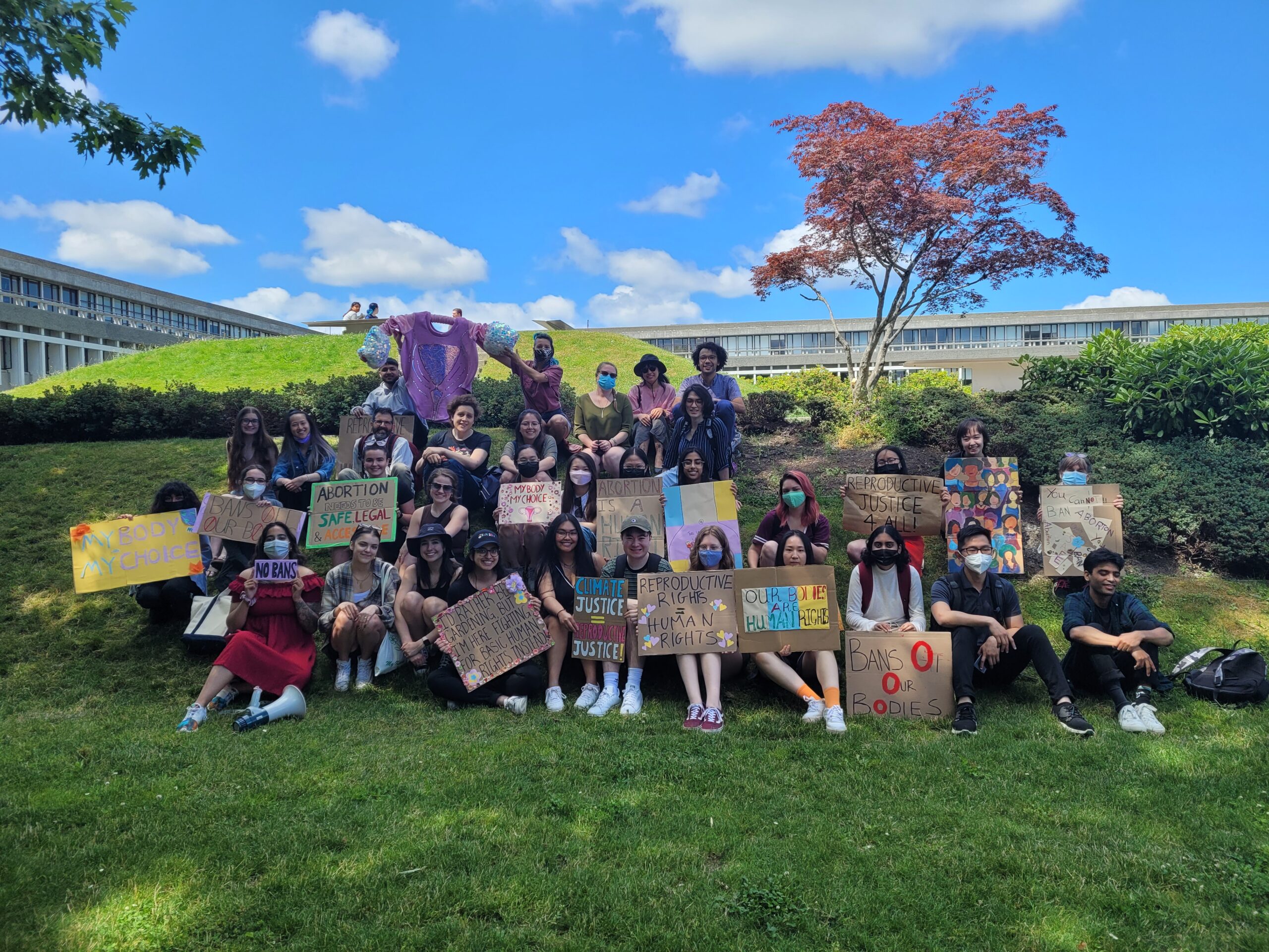 Roughly 35 students sit on the grassy hill at the SFU Burnaby Campus. They are collected, smiling for the camera. They hold signs that read, “No Bans,” Reproductive Justice for all,” “Reproductive Rights are human rights,” and more.