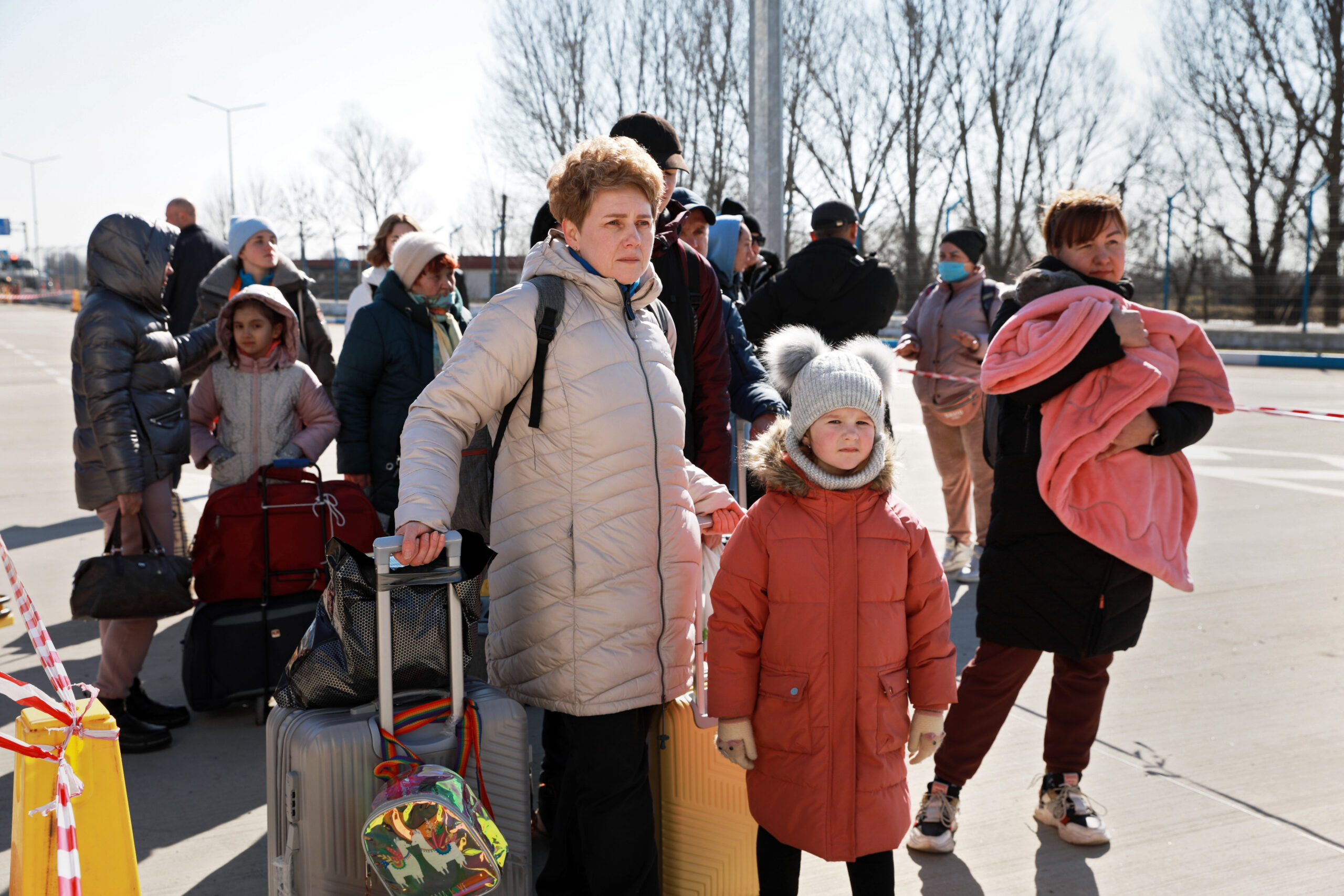 The photo is of Ukrainian people fleeing from their country. In the centre of the photo is a women with her arm around a small child. They are holding multiple bags of luggage and are surrounded by others with more luggage.