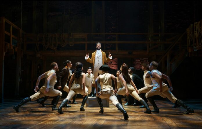 Hamilton_JoanMarcus A standing Hamilton is surrounded by the ensemble who are in a lunching position towards him