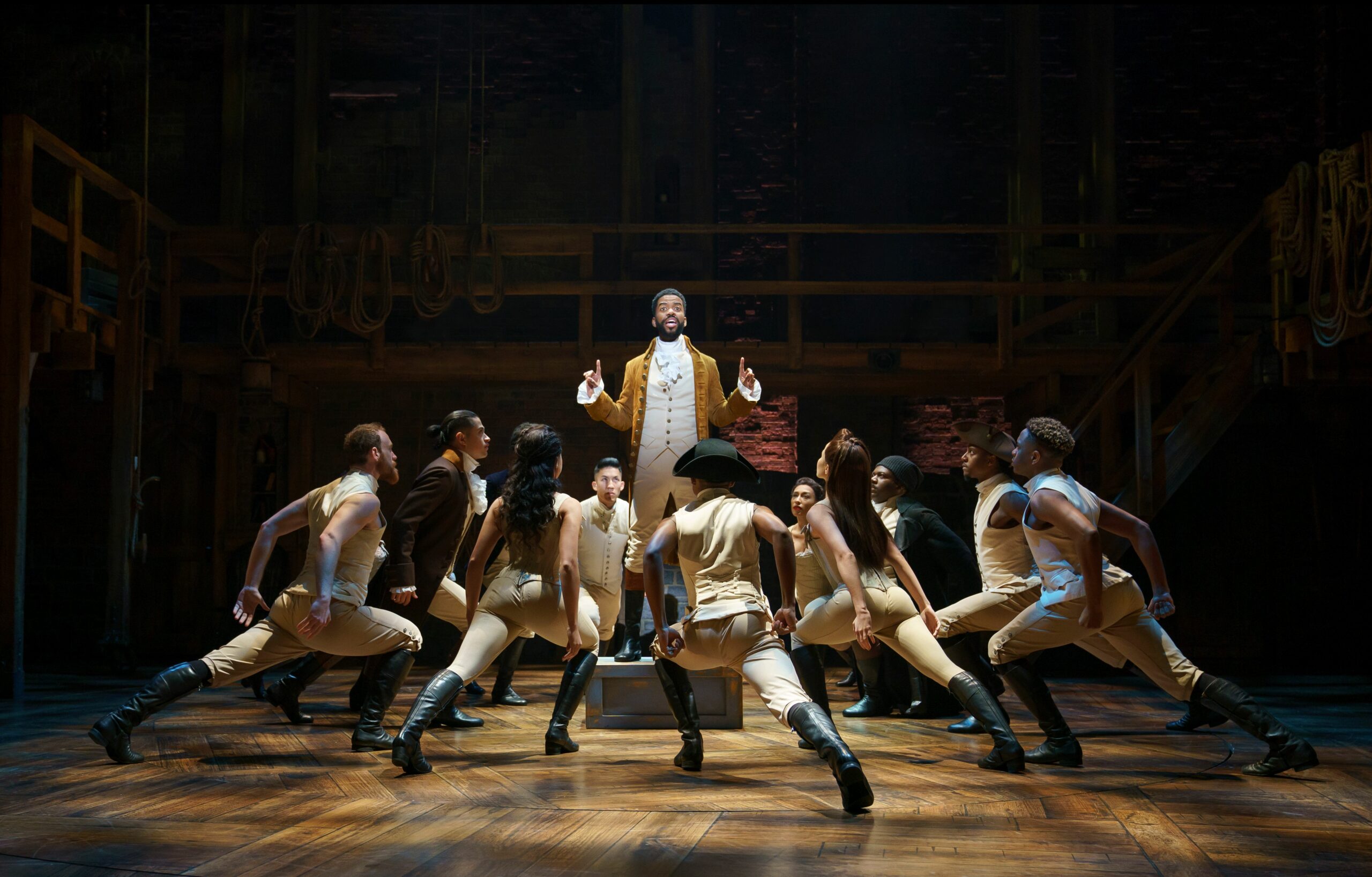 A standing Hamilton is surrounded by the ensemble who are in a lunching position towards him