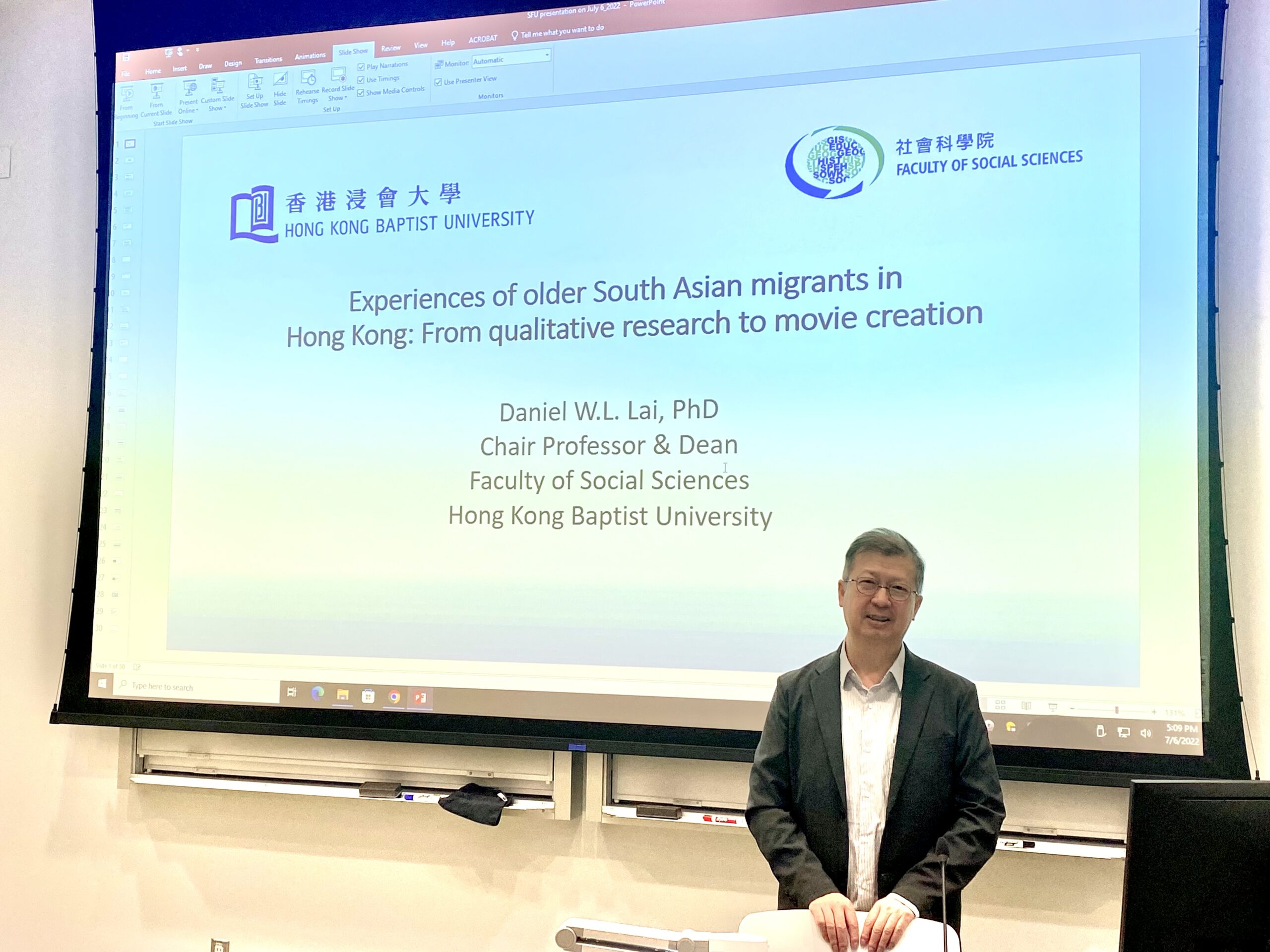 Professor Lai is standing in front of a projector screen that reads: Experiences of older South Asain migrants in Hong Kong: From qualitative research to movie creation. He is looking at the camera smiling.