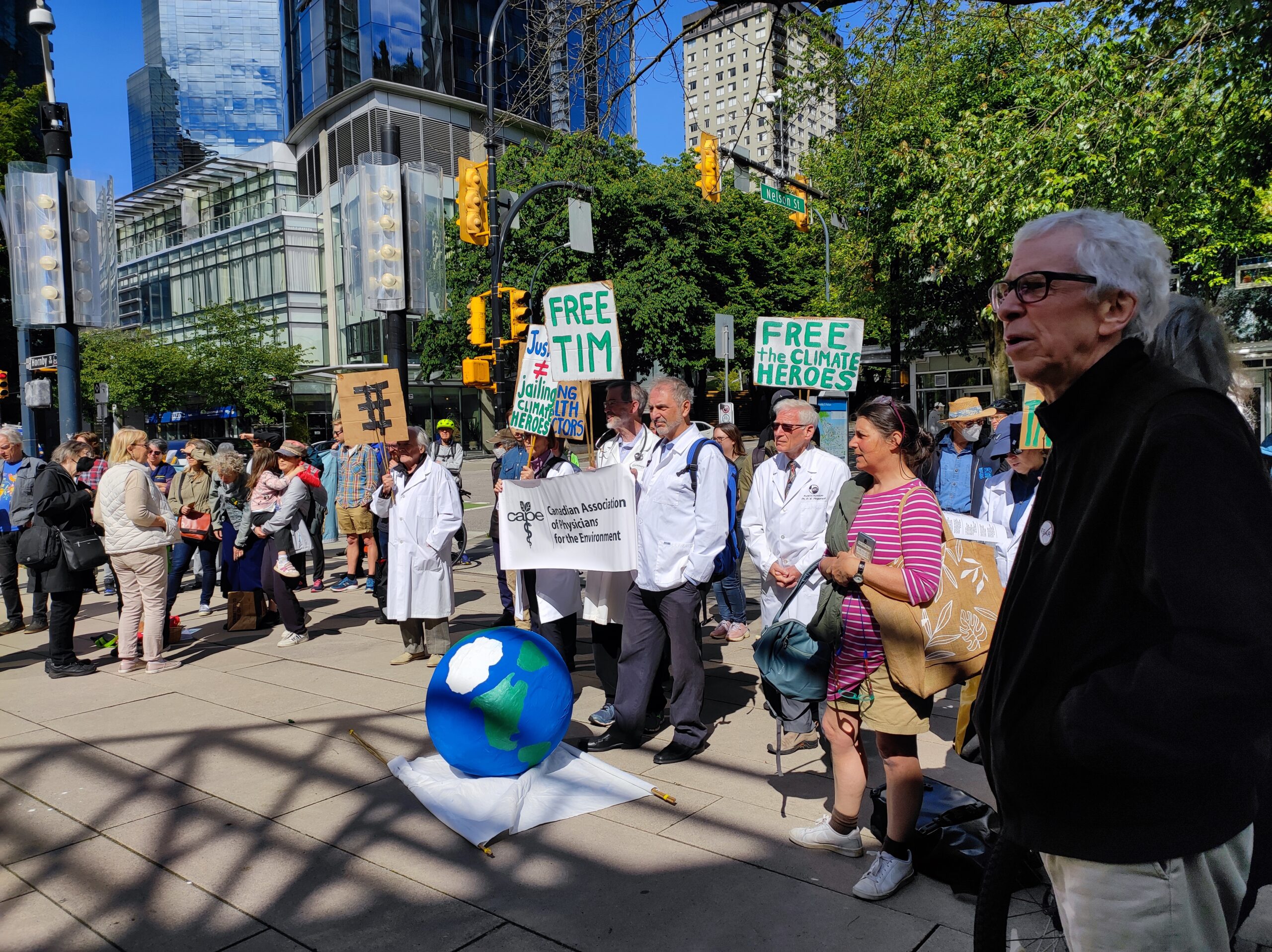 The photo is of a large crowd, gathered outside in Vancouver. They are holding signs that read, “Free Tim” and “Free the climate heroes.” Two people are holding a large banner that says “Canadian Association of Physicians for the Environment.”
