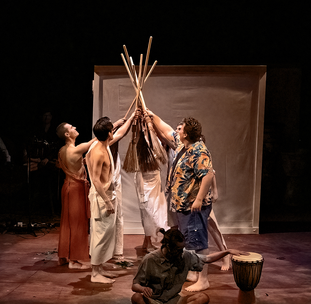 A group of white men holding sticks and pointing them together facing each other, with one man at the front playing a drum