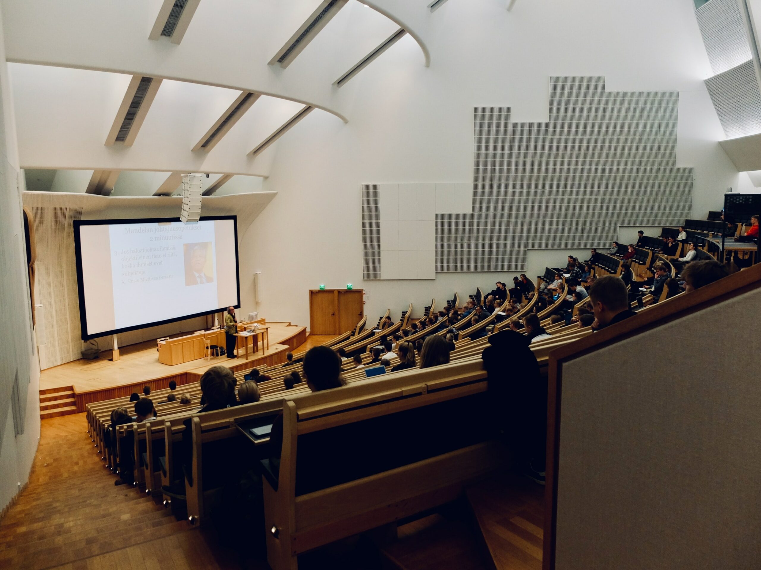 The photo is of the inside of a lecture hall. The lecture hall is full of students, attention turned to the front of the room where the professor is delivering a lecture presentation.