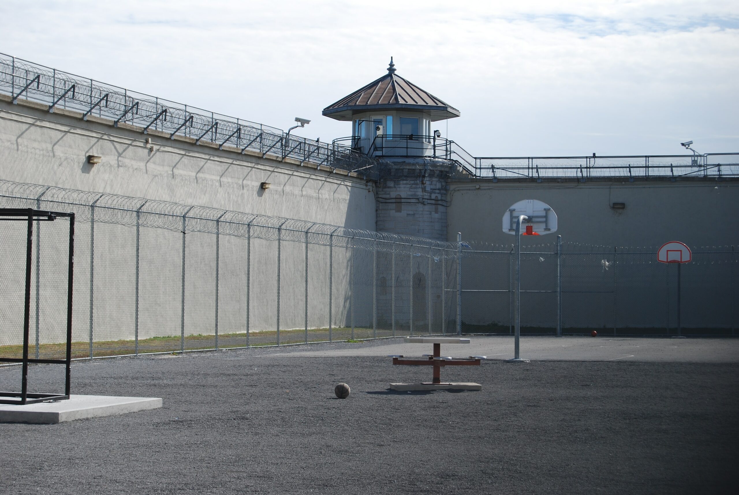 The photo is of the outside of a Canadian prison. A basketball sits in front of a basketball hoop in a gravel field, surrounded by fencing. No people can be seen.