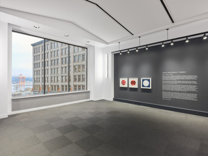 paintings in square frames are lined up against a blank wall, pictured is a large window on another wall over looking the waterfront