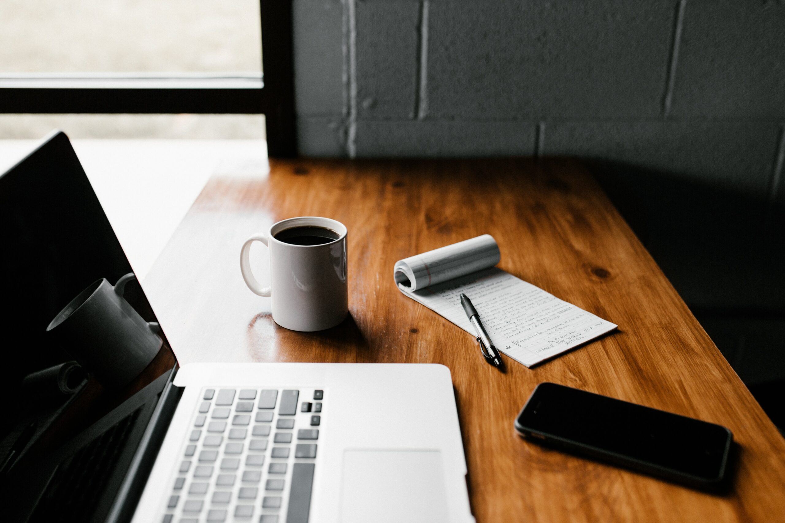 a laptop, cup of coffee, notebook and pen, and phone on table