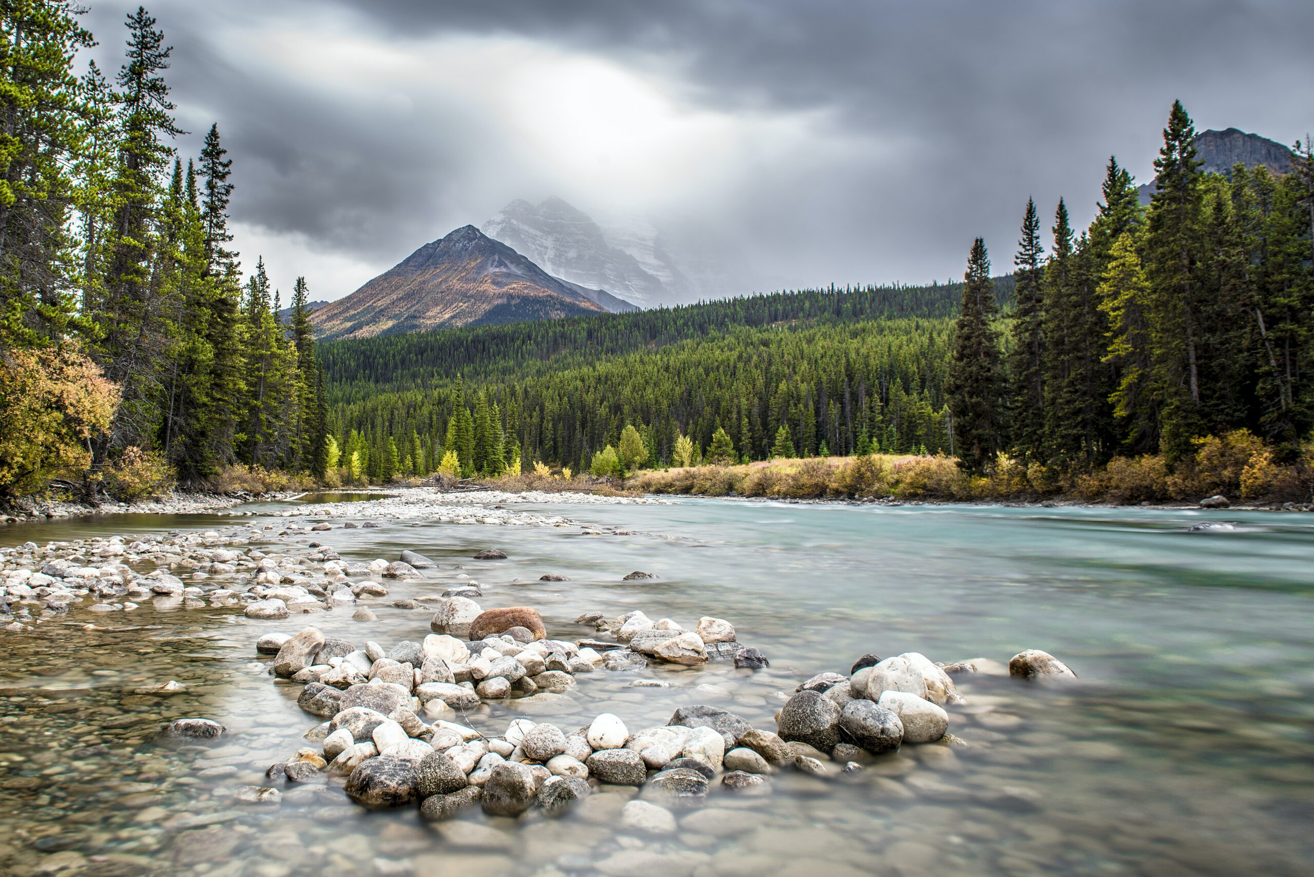 The photo is of the Canadian outdoors. Large mountains are seen in the background. A river runs through a green forest.